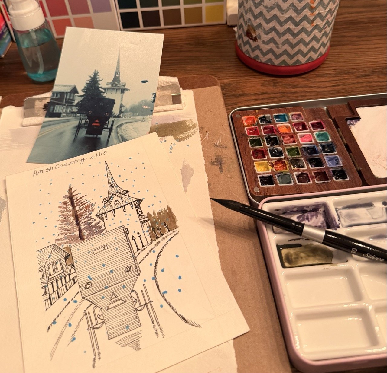 An art workspace with watercolor paints and brushes, featuring a hand-drawn sketch and a photographic reference of a church with a tall steeple and trees, labeled 'Amish Country Ohio'.