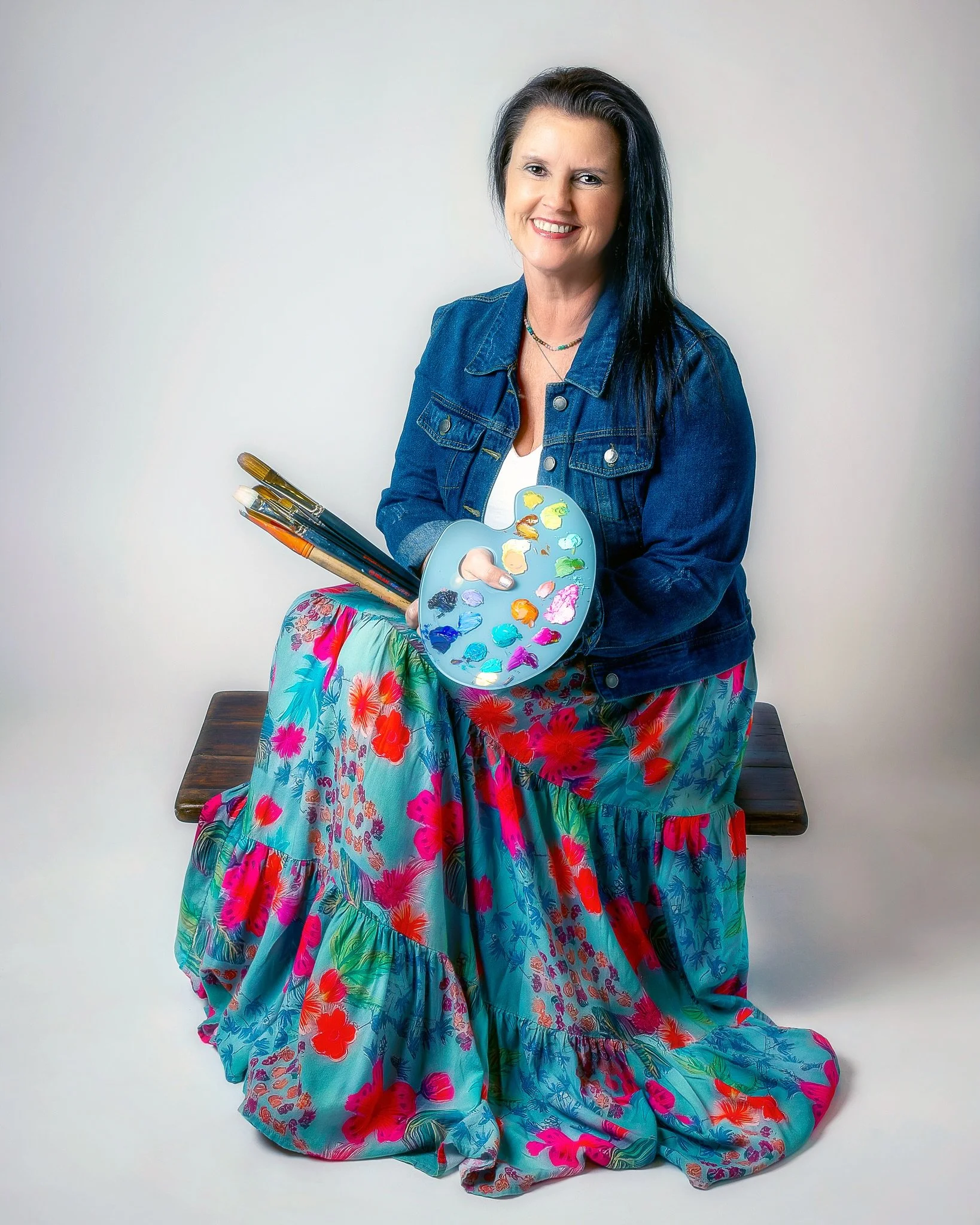 A woman with long black hair, smiling, dressed in a denim jacket, holding a painter's palette and brushes, seated on a bench with a colorful floral skirt, against a plain white background.