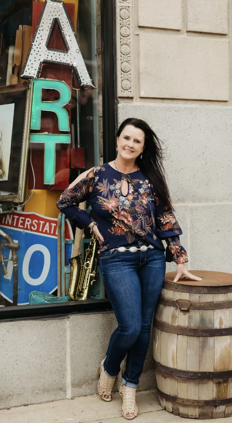 A woman with long dark hair standing outside a vintage store with neon signs, wearing a floral blouse, jeans, and beige high-heeled boots, leaning on a wooden barrel.