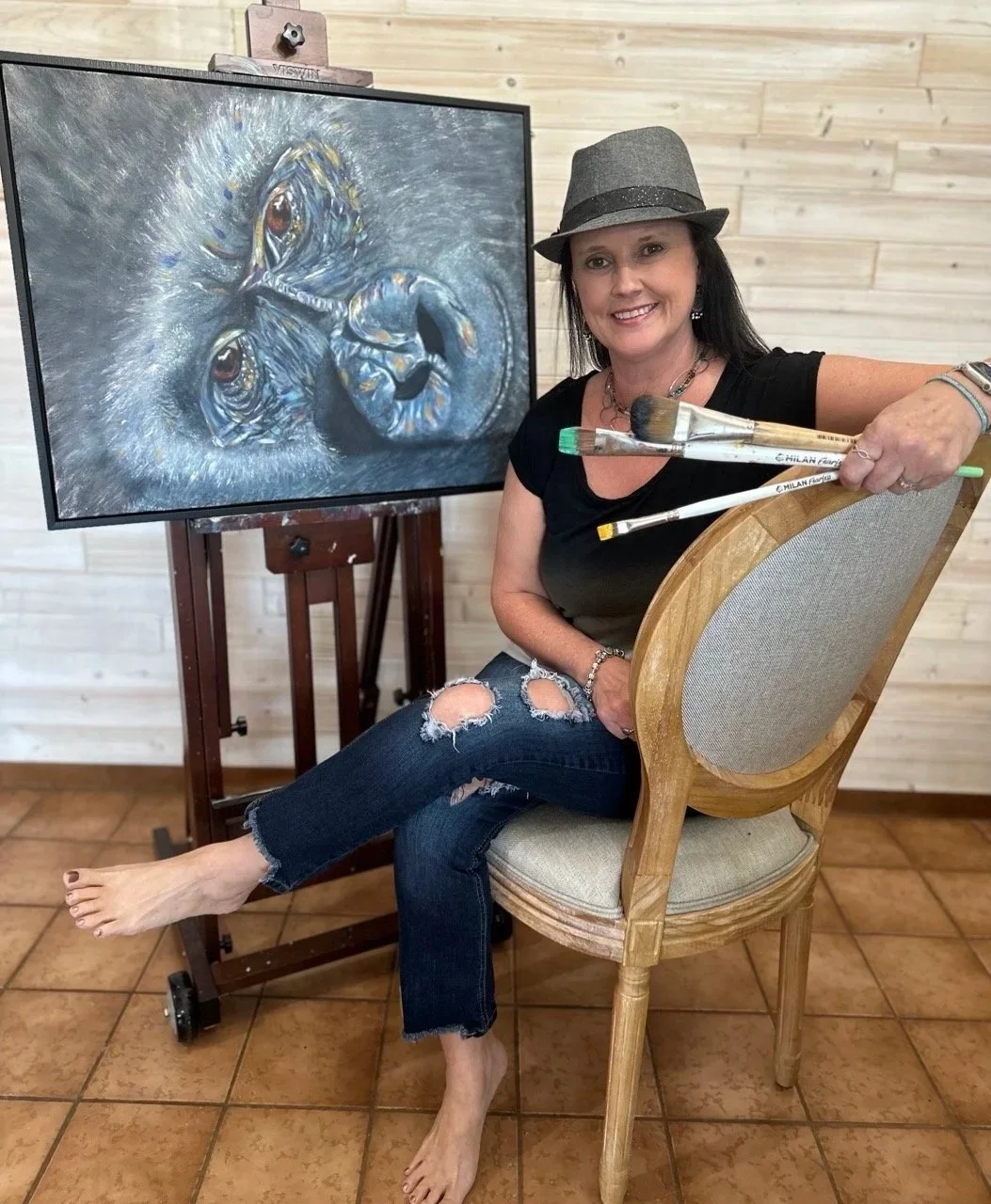 A woman sitting barefoot on a wooden chair holding paintbrushes, smiling next to a realistic lion painting on an easel, with a light wooden wall in the background.