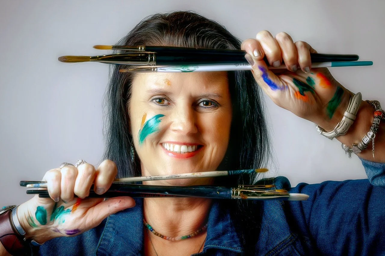 A woman with dark hair smiling, holding paintbrushes in front of her face, with colorful paint strokes on her cheeks and hands, wearing a blue jacket and a beaded necklace.