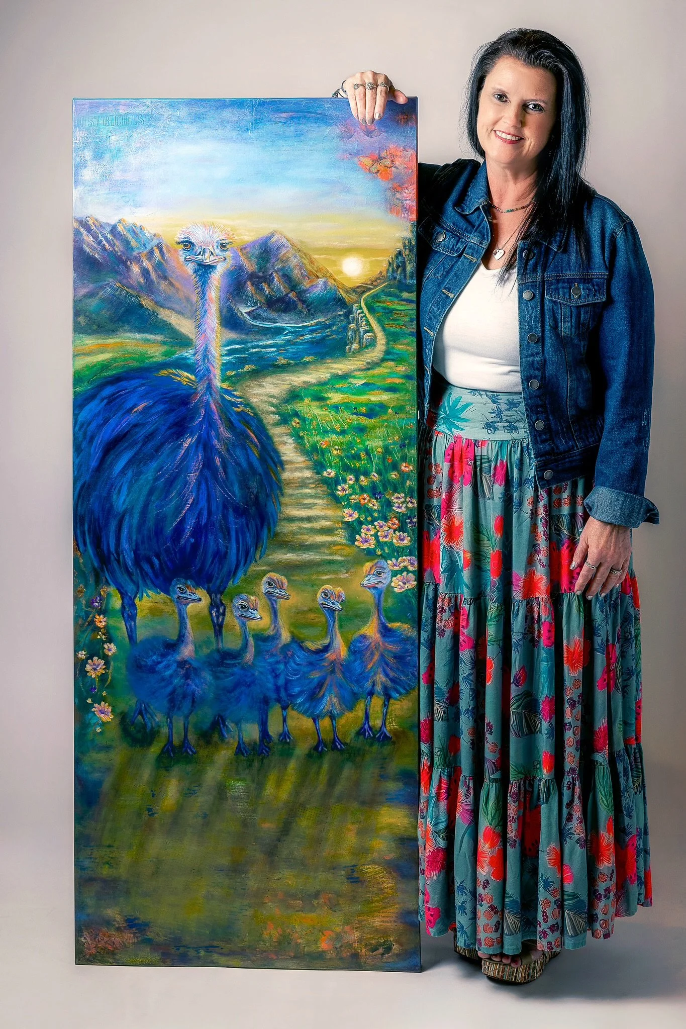 A woman with black hair, wearing a denim jacket, white top, and colorful long skirt, standing next to a large colorful painting of a landscape with a large blue bird, mountains, a sunset, and several smaller birds.