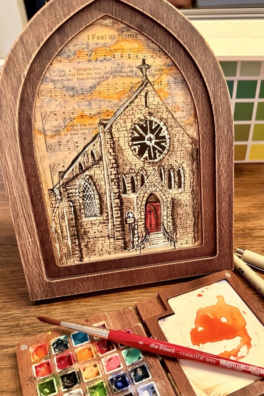 A watercolor painting of a small, old brick church with a red door and a large round stained glass window, set inside a wooden frame. The background features a sheet of music and some text that reads 'I Feel at Home.'