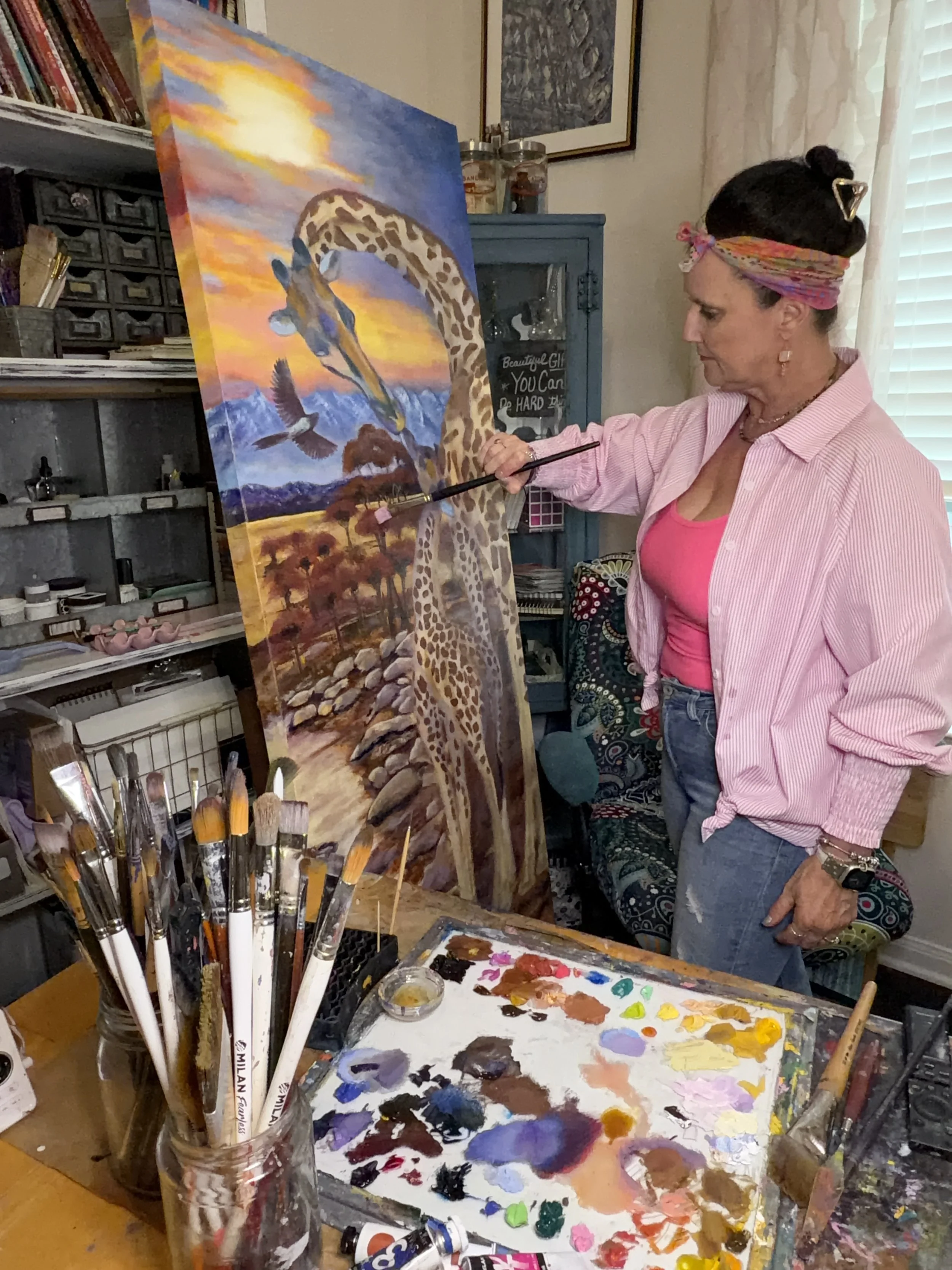 An artist wearing a pink shirt and a colorful headband is painting a sunset landscape with a giraffe and mountains on a canvas. Art supplies including paintbrushes, paints, and a palette are visible on the table.