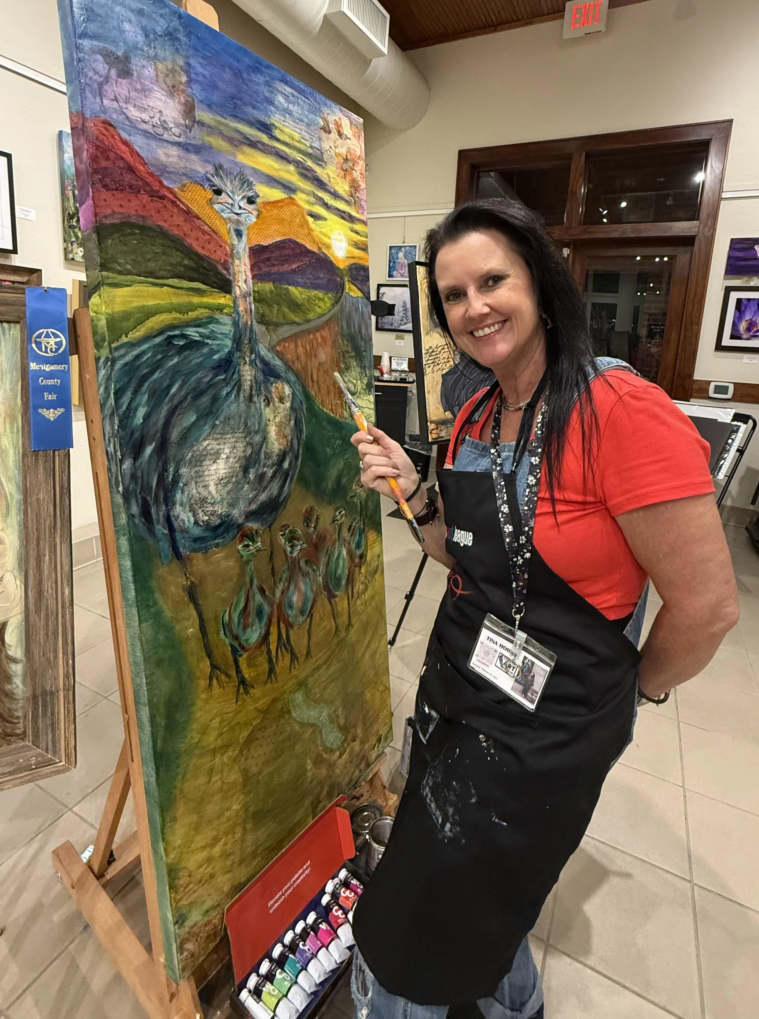 A woman smiling, holding a paintbrush, standing next to a colorful abstract landscape painting of a bird on a easel in an art gallery. She wears a red shirt, black apron, and jeans, with art supplies on the floor.