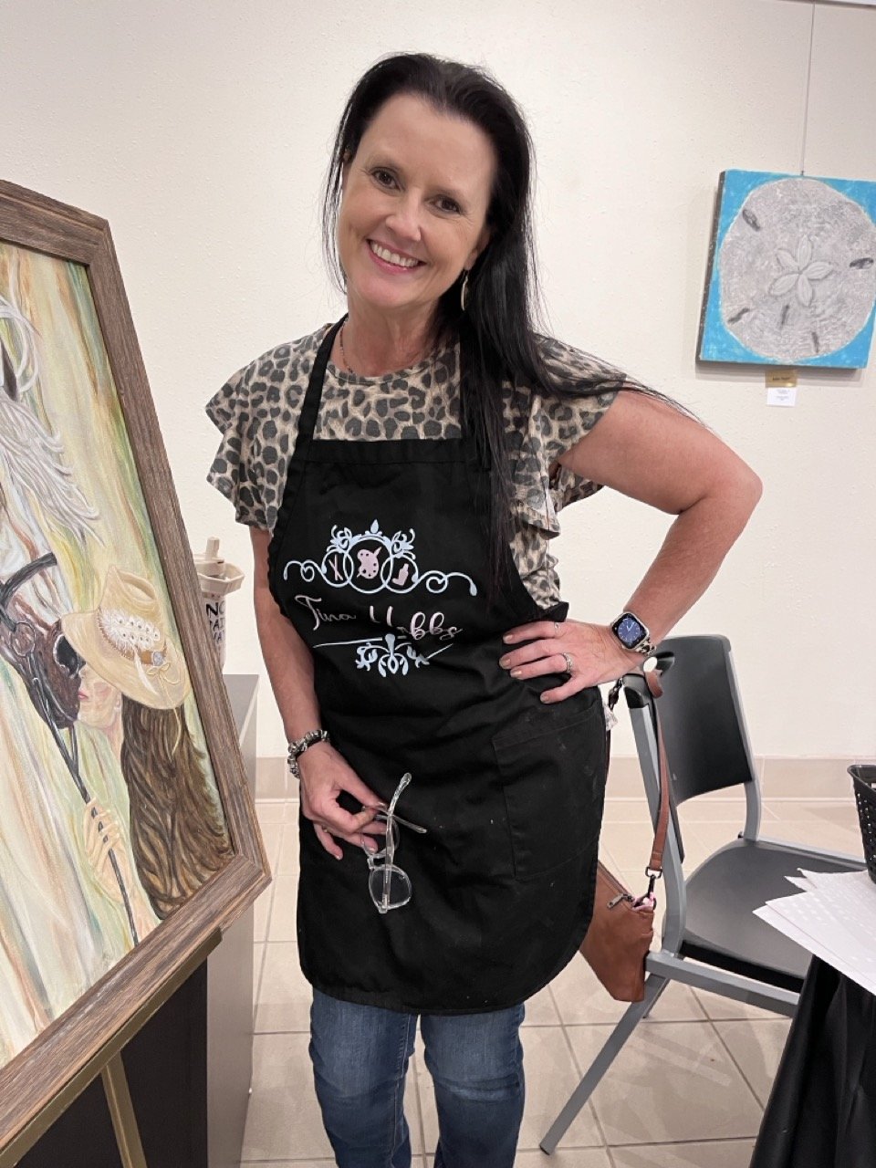 A woman with dark hair wearing a black apron and leopard print top, holding glasses, smiling, standing next to artwork in an art gallery or studio.