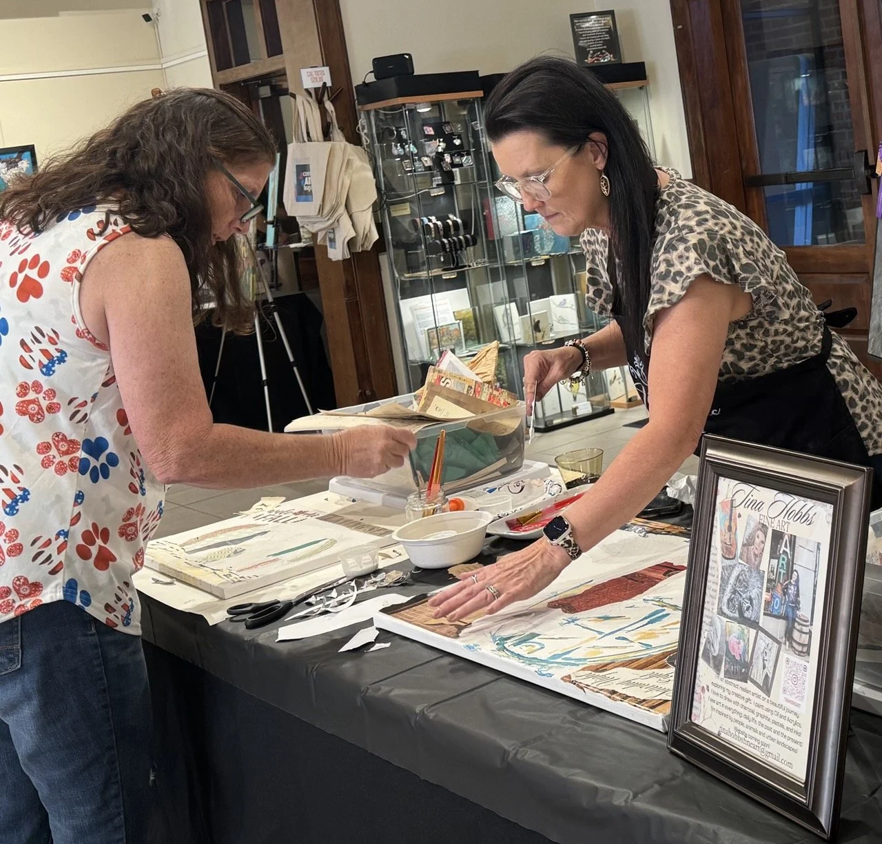 Two women working on an art project at a table indoors, with art supplies and framed artwork visible.