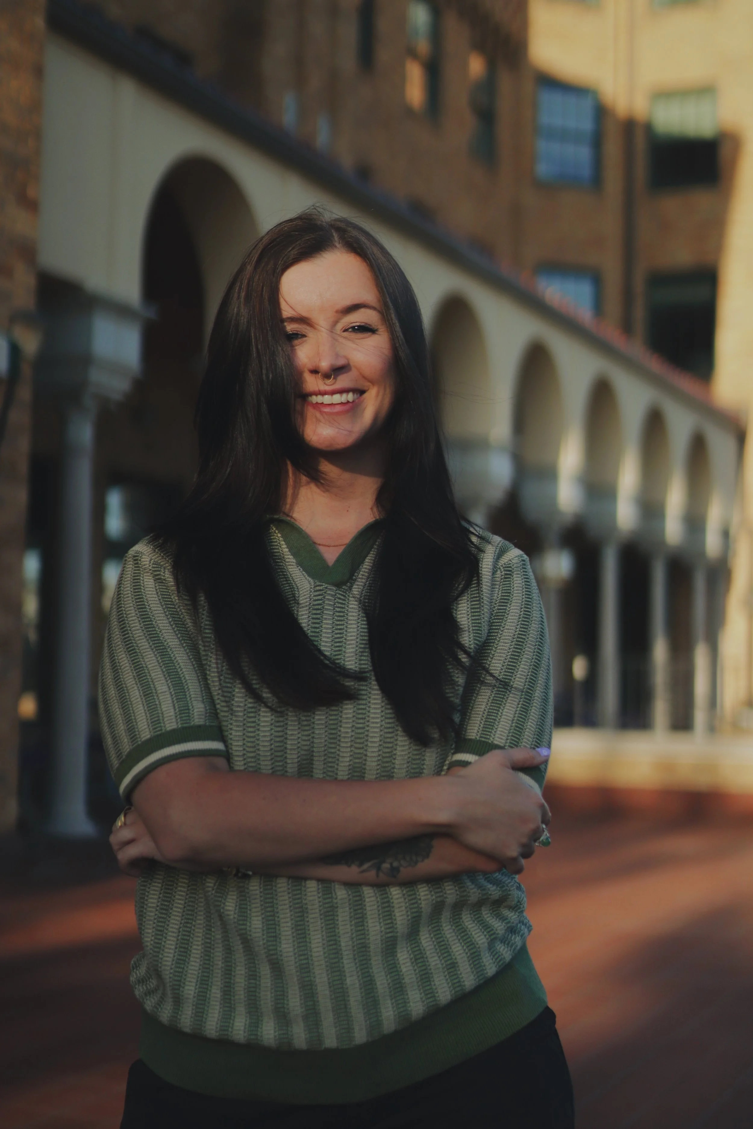 A woman with long dark hair smiling, wearing a patterned green and gray short-sleeve shirt, with her arms crossed, standing outdoors in front of a row of arches and a brick building during sunset.