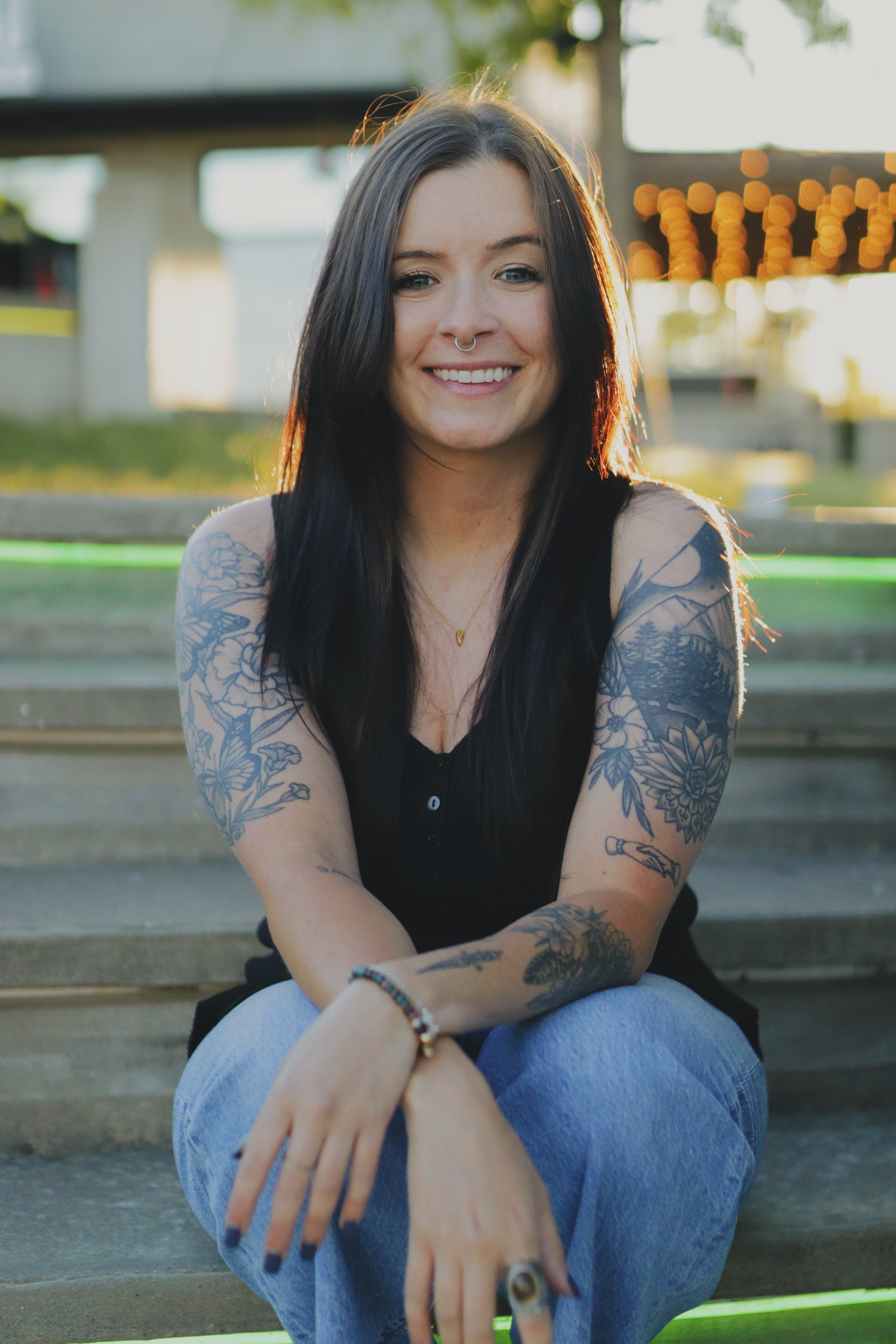 A young woman with long dark hair, tattoos on her arms, and a septum piercing, sitting outdoors on steps at sunset, smiling at the camera.