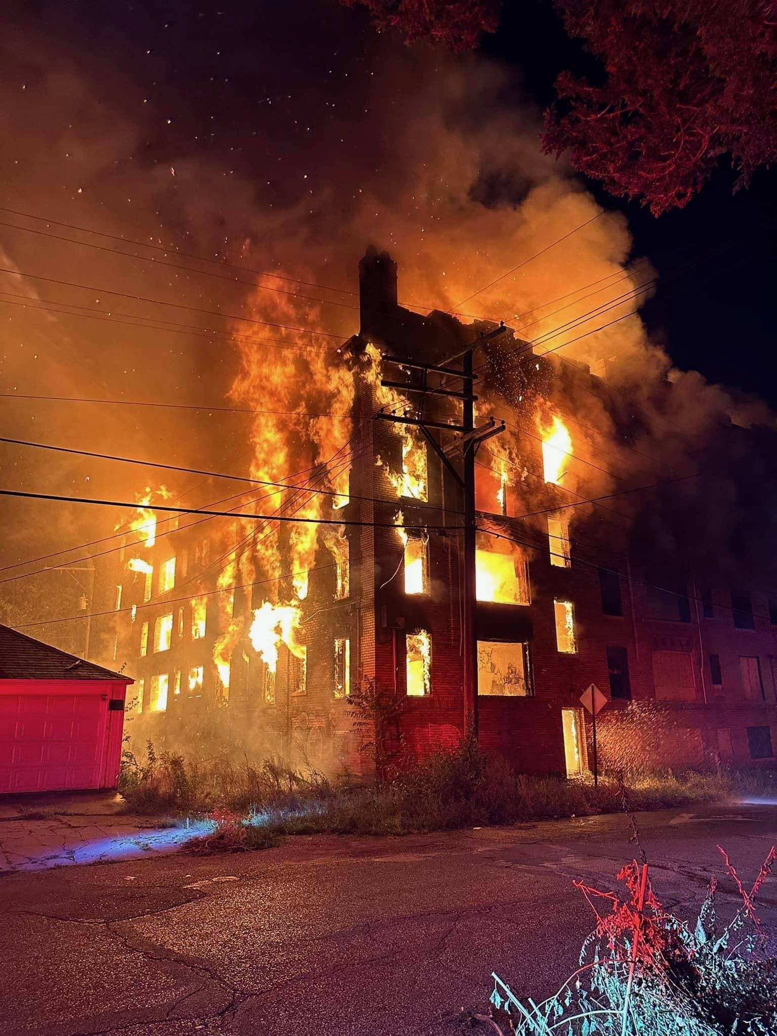 A house fire with flames engulfing an entire multi-story building at night, smoke billowing into the sky, and utility poles and wires in the foreground.