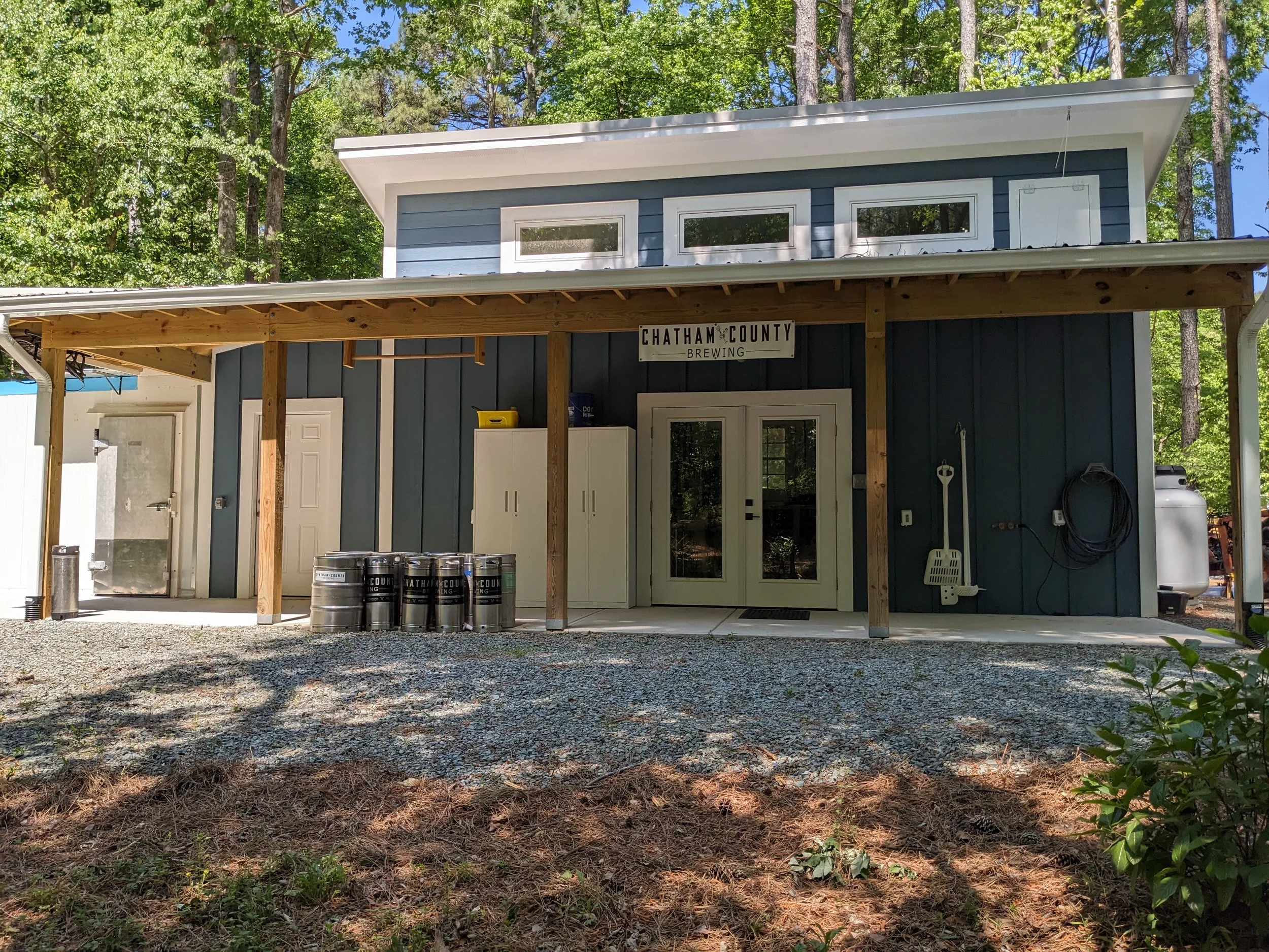 A two-story building with dark blue horizontal siding, white trim, and a slanted roof, situated among tall trees with green leaves. The front of the building features a sign that reads "Chatham County Brewing" and has three small rectangular windows on the upper floor. The lower section has a closed door, cabinets, a propane tank, and some barrels in front. The ground is covered with gravel and some patches of grass.