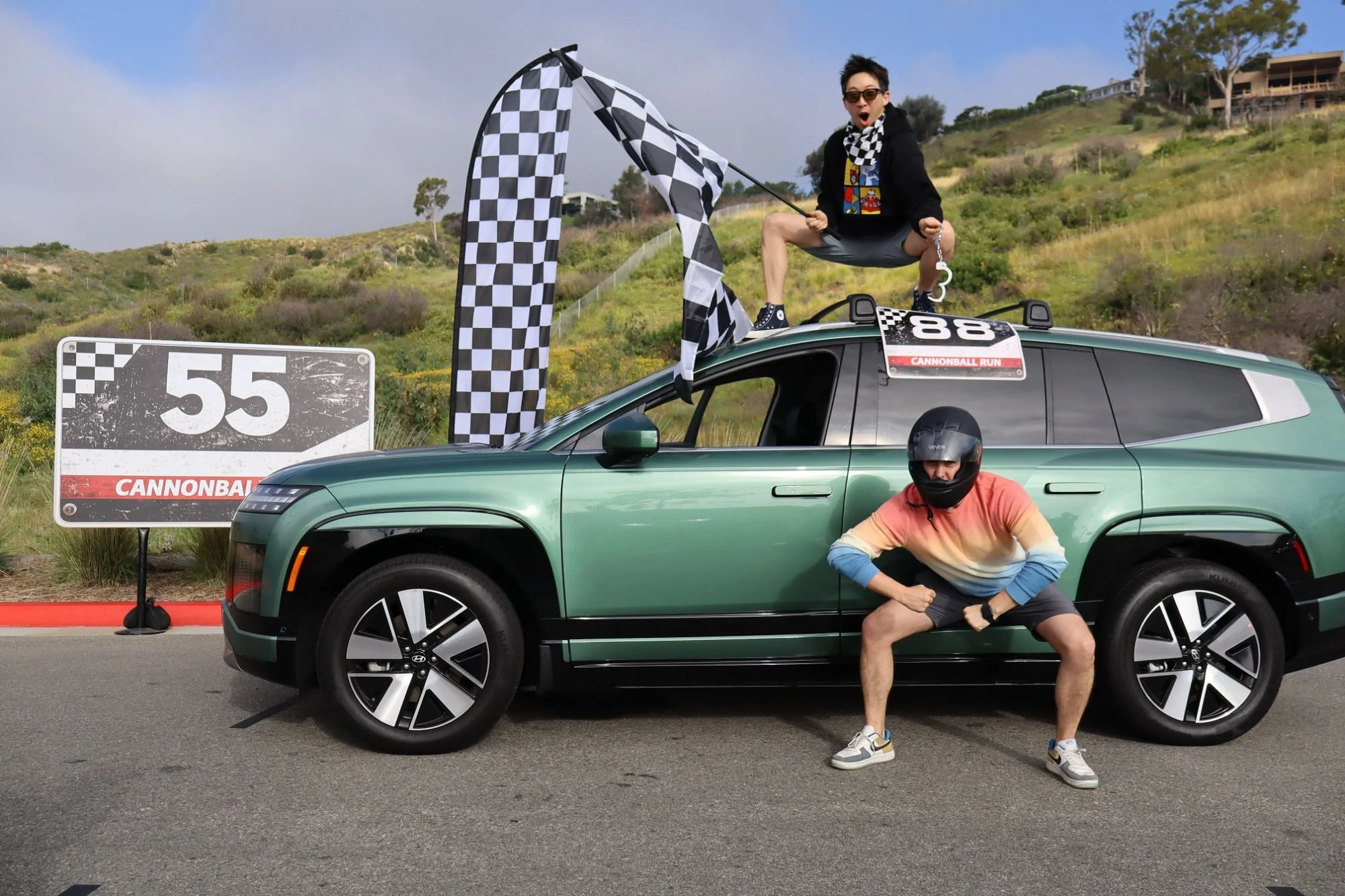 A person standing on top of a green car, holding a checkered flag and making an excited expression, with another person squatting below the car wearing a helmet and making a fist gesture. The background features a hillside with trees and houses, and a sign that reads '55 CANNONBALL' and a checkered flag pattern, along with a small sign that says 'CANNONBALL RUN'.