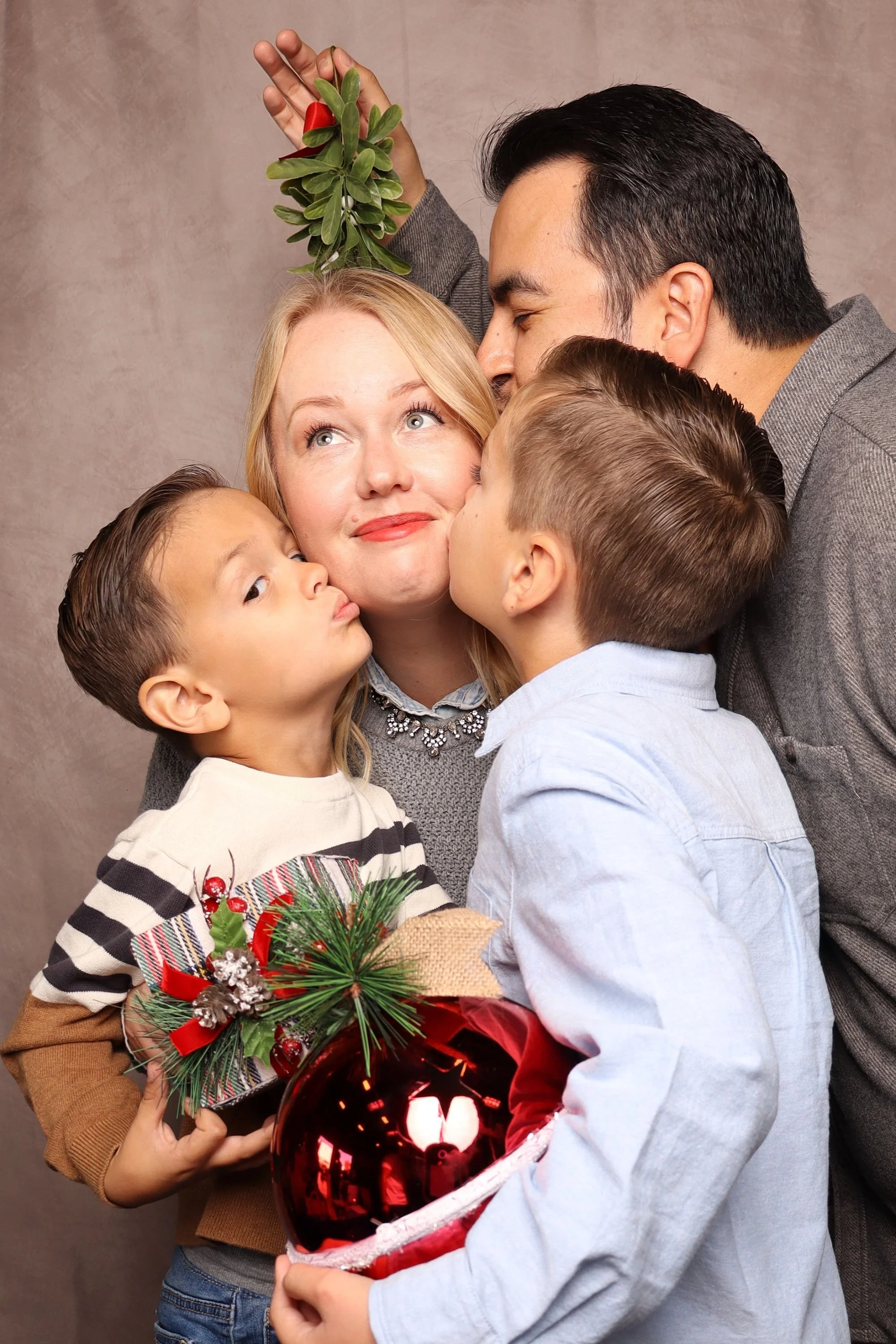 A family gathered for Christmas, with children and adults sharing a moment of affection, holding a Christmas ornament shaped like a heart with a snow globe scene inside, decorated with pine branches, berries, and a red bow, against a neutral background.