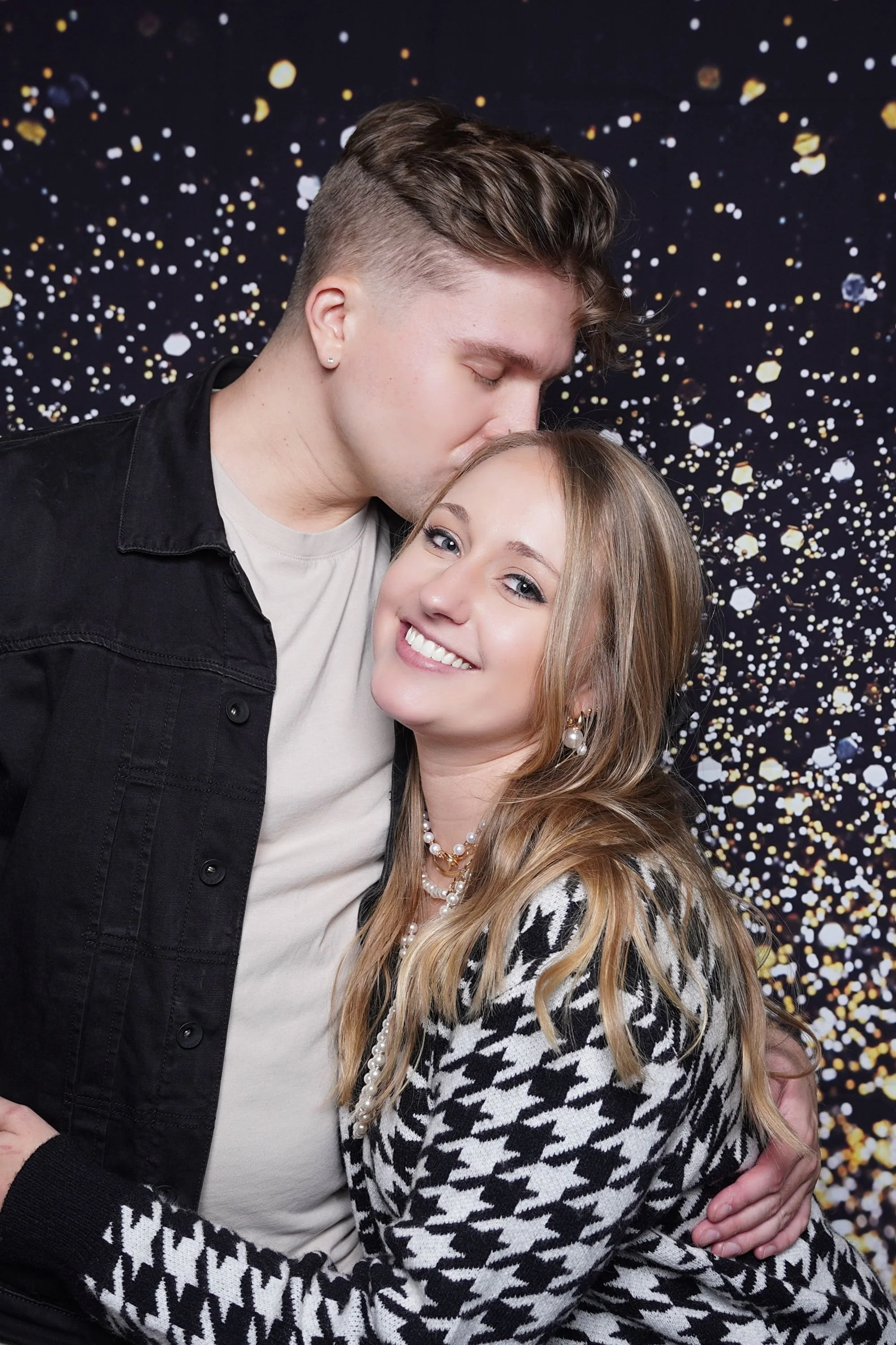 A young man with short, styled hair and earrings kisses a smiling young woman with wavy hair, wearing pearl jewelry, against a backdrop of black with gold, white, and silver confetti-like spots.