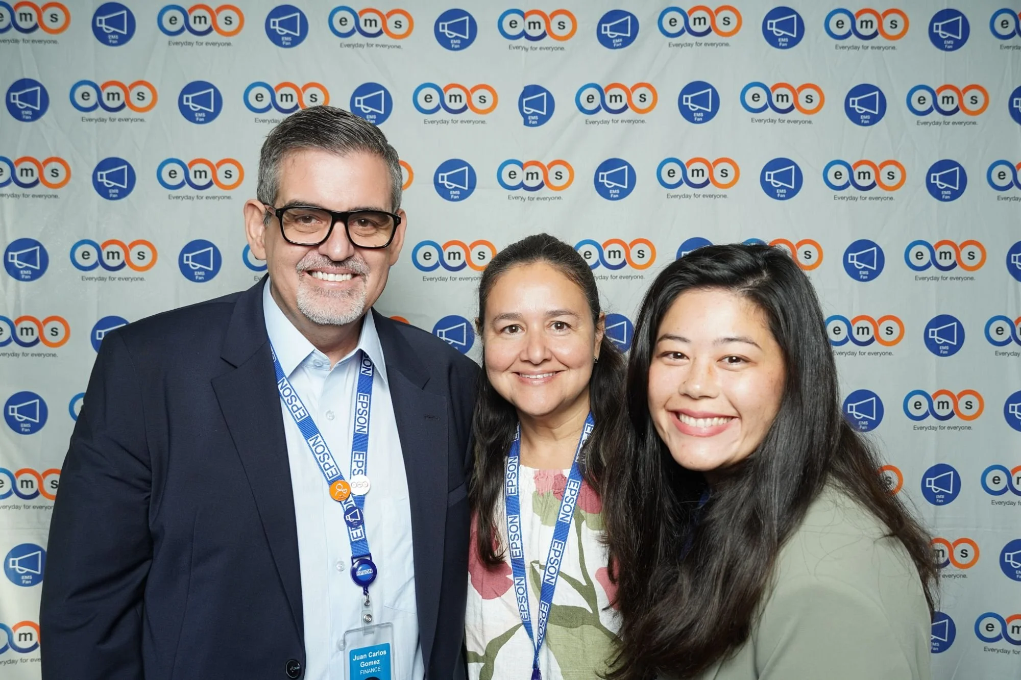 Three people smiling in front of a backdrop with the EMS logo and slogan 'Everyday for everyone.' The man on the left wears glasses and a suit, the woman in the middle has dark hair and is wearing a light-colored top with a floral pattern, and the woman on the right has long dark hair with loose waves.