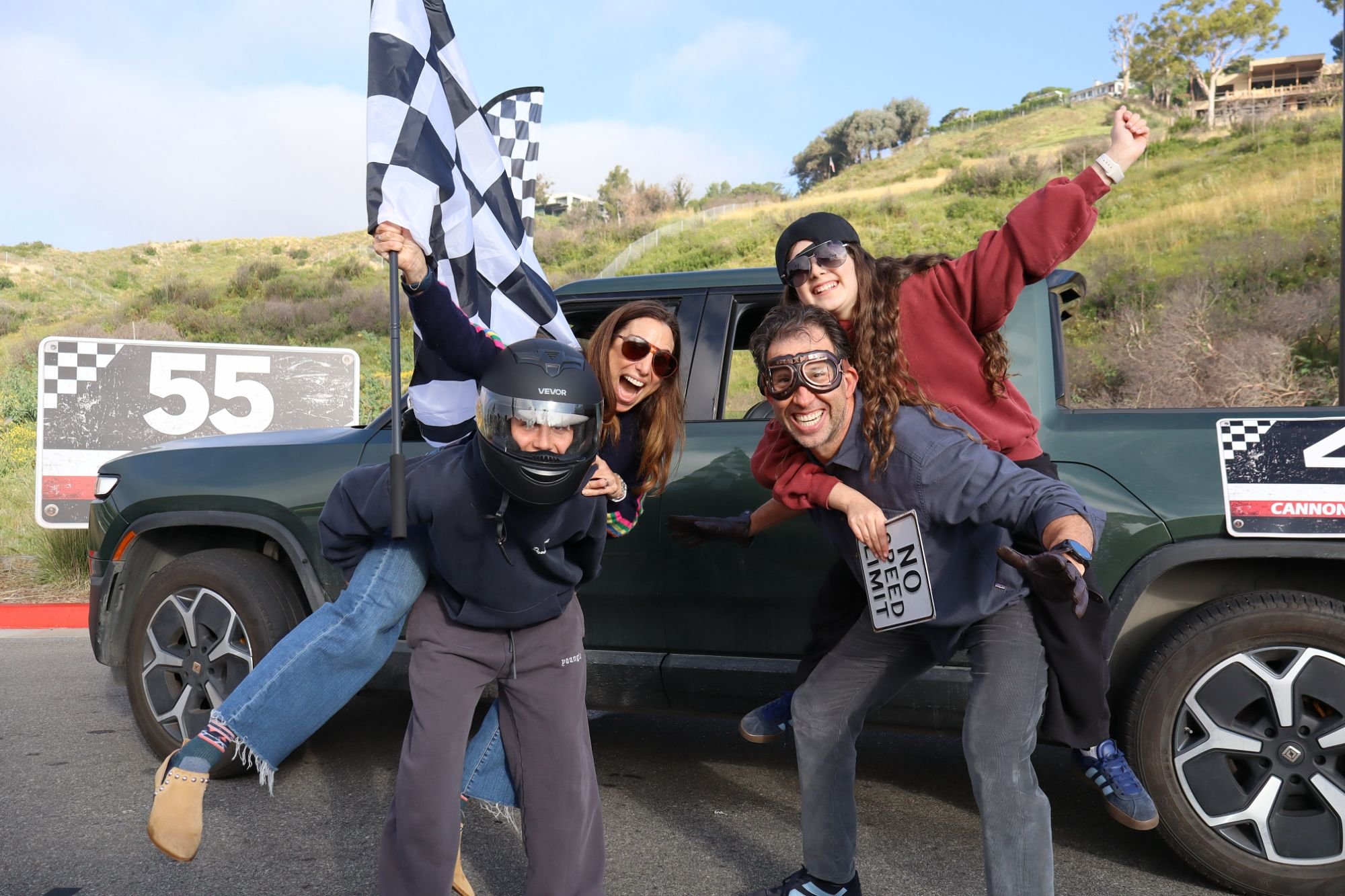 Group of people celebrating at a racing event, holding a checkered flag, posing in front of a green car, with hills and houses in the background.