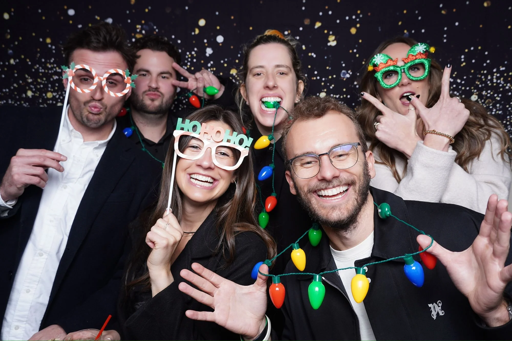 A group of six friends celebrating at a holiday party, wearing Christmas-themed glasses, shiny necklaces, and holiday lights, with a starry background.