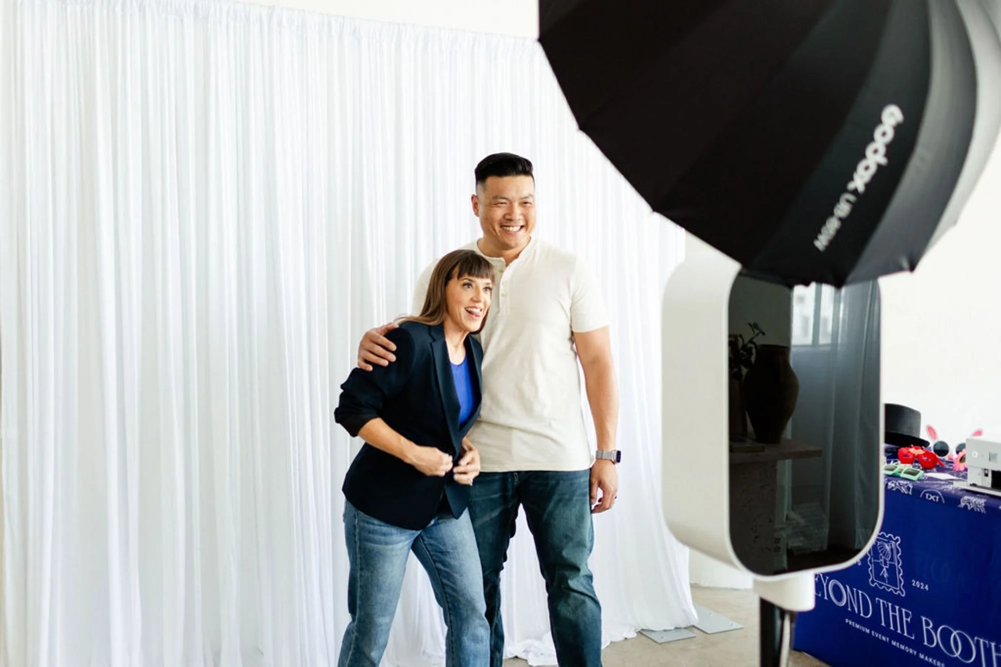 A man and a woman taking a photo with a smartphone in front of a backdrop. The man is smiling and wearing a white shirt, and the woman is also smiling, dressed in a blue blazer with jeans. There is photography equipment such as a large umbrella light in the foreground, and a table with various items in the background.