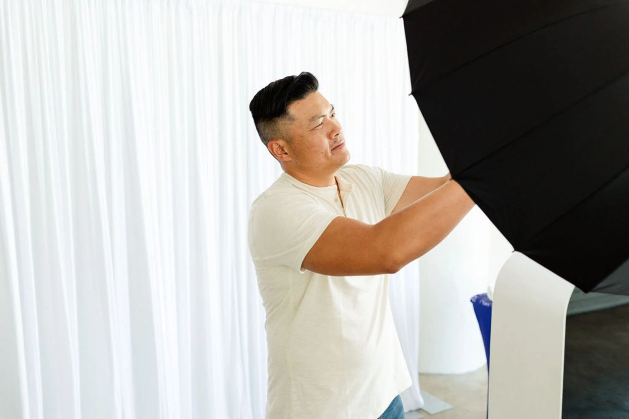 A man with dark hair wearing a white shirt is adjusting a large black umbrella or softbox on a photography setup in a bright room with white vertical blinds.