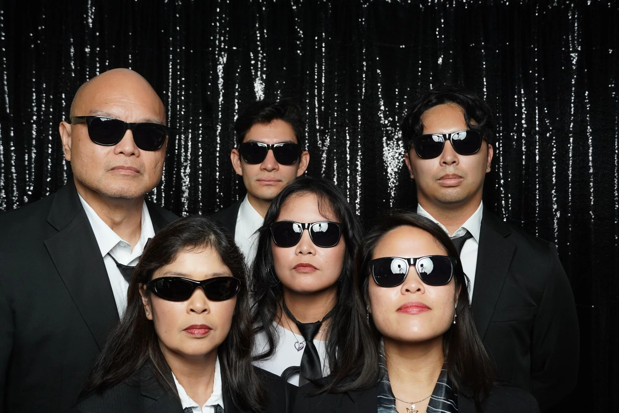 Group of people wearing black suits and black sunglasses standing in front of a glittery black backdrop.