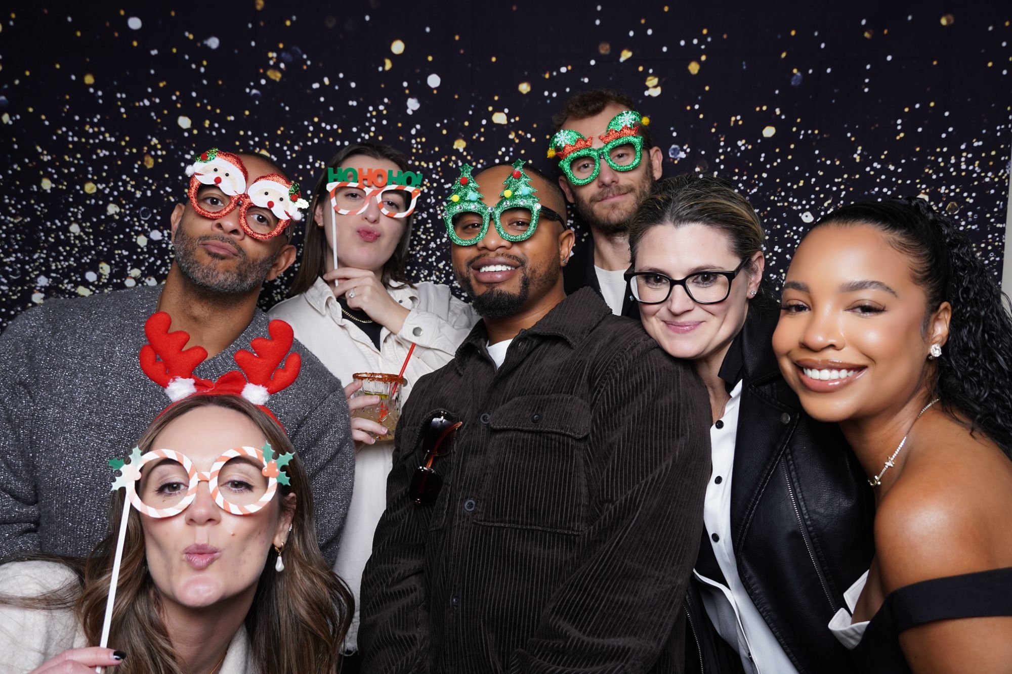 A group of six diverse people at a festive party wearing holiday-themed novelty glasses and accessories, with a starry night backdrop.