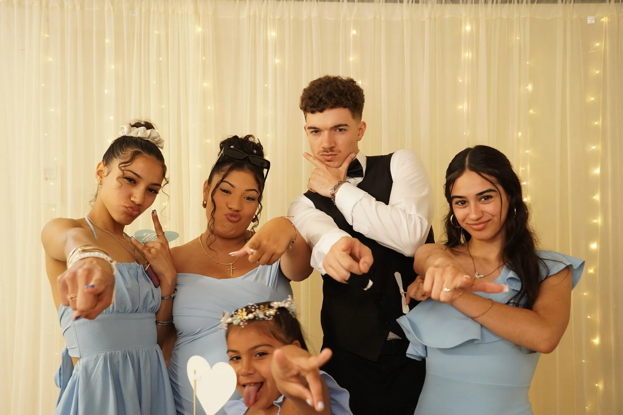 Group of five young people posing together at a celebration, all pointing towards the camera. They are dressed nicely, with three females wearing light blue dresses and one male in a tuxedo. The background features a cream-colored curtain decorated with string lights, creating a festive atmosphere.