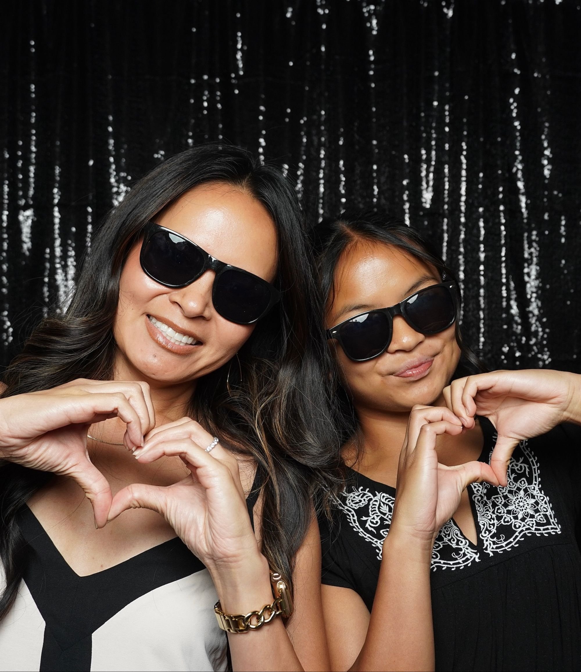 Two smiling women wearing sunglasses and holding hands in a heart shape, posing against a sparkling black backdrop.