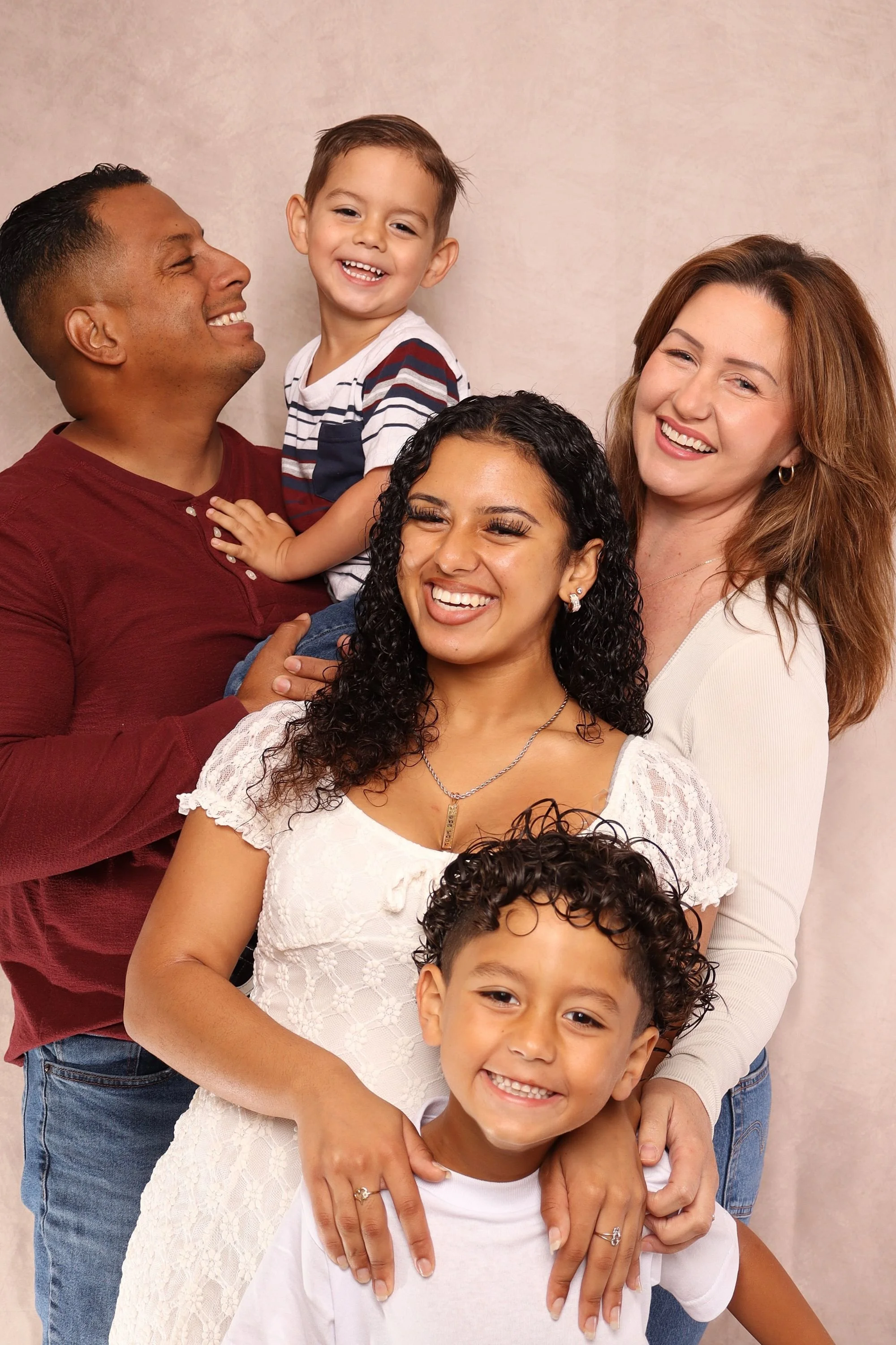 A happy multiracial family of five posing together against a neutral background. Two adults and three children, smiling and embracing each other.