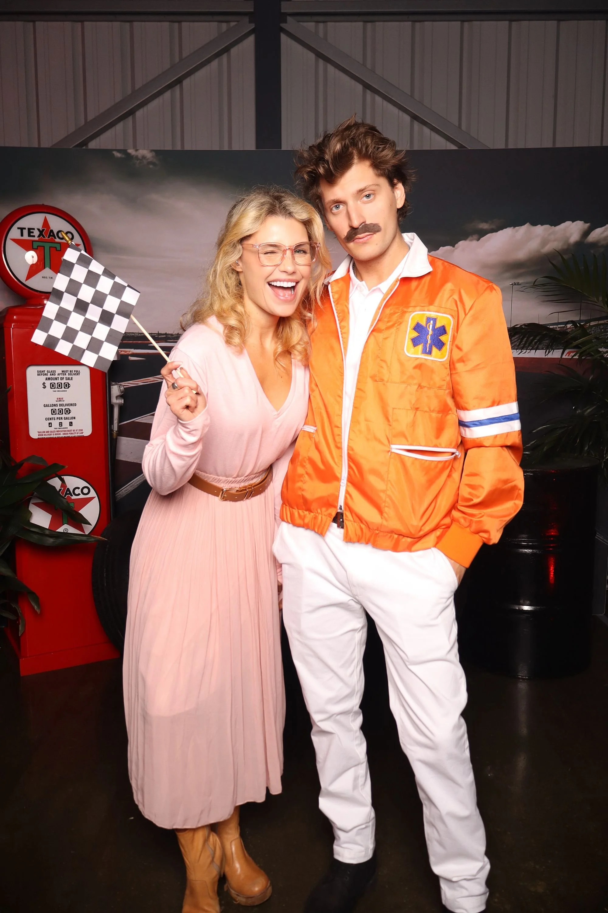 A woman in a light pink dress and brown boots smiling and holding a checkered flag, standing next to a man in a bright orange rescue worker jacket with a fake mustache, in front of a vintage Texaco gas station backdrop.