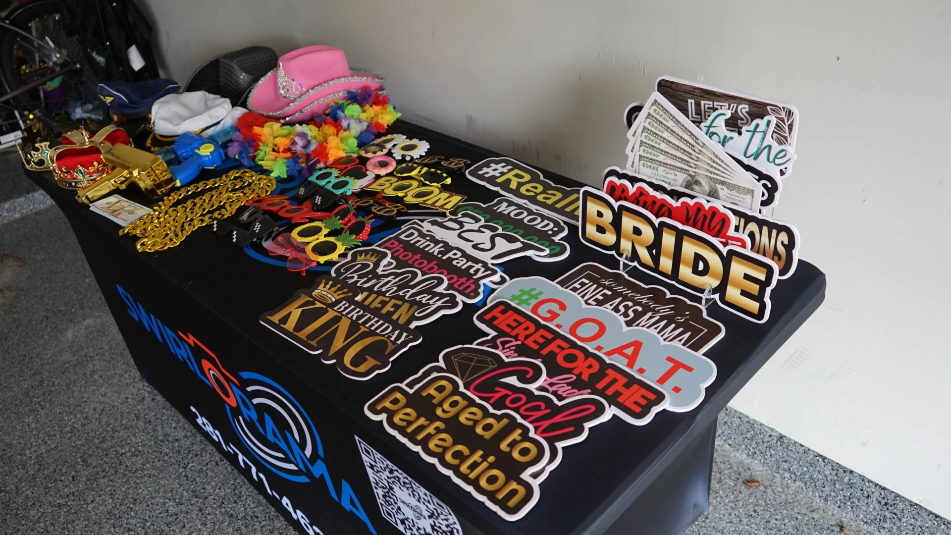 Table displaying various photo booth props including oversized sunglasses, feathered headbands, flower leis, tiaras, and signs with messages like "BRIDE," "Groom," "HAPPY BIRTHDAY," and "Let's for the." In the background, there is a pink tiara and a pink hat with a tiara on top.