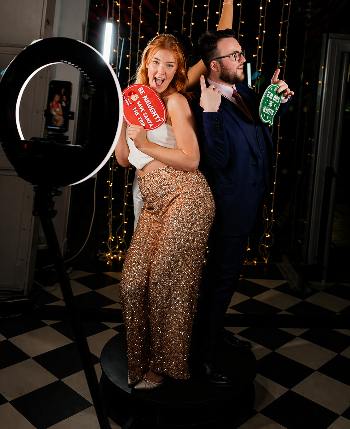 A joyful man and woman posing on a photo booth platform at a celebration. The woman, with red hair, is wearing a white top and shiny gold pants, holding a red sign that says "Be Naughty Save Santa The Trip." The man, wearing glasses and a dark suit, holds a green sign that says "I'M ONLY A MORNING PERSON ONCE A YEAR." There are fairy lights in the background and a ring light illuminating the scene.