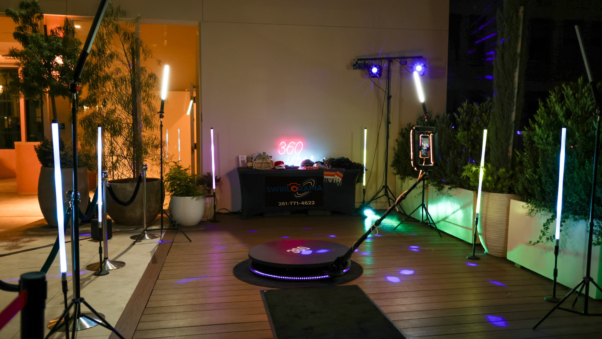 Indoor event space with DJ setup, colorful vertical LED lights, a round dance platform, and potted plants, illuminated by decorative lighting.