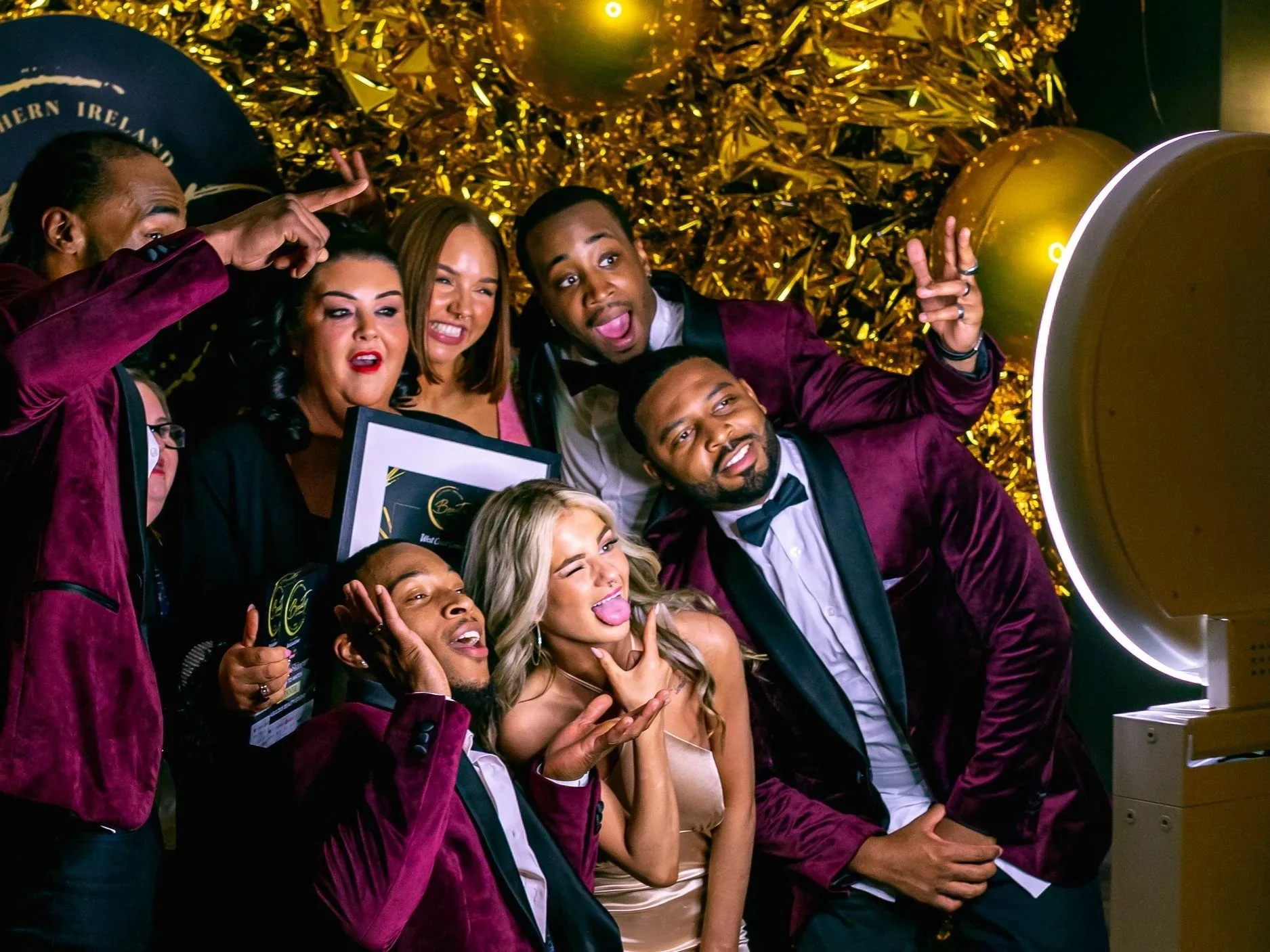 A group of people dressed in formal attire are taking a selfie together in front of a gold and black backdrop with balloons. They are posing playfully with smiles and gestures, celebrating a special event or party.