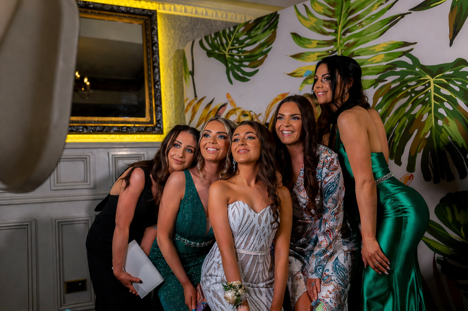 Group of five women in formal attire taking a selfie together at a decorated event venue.