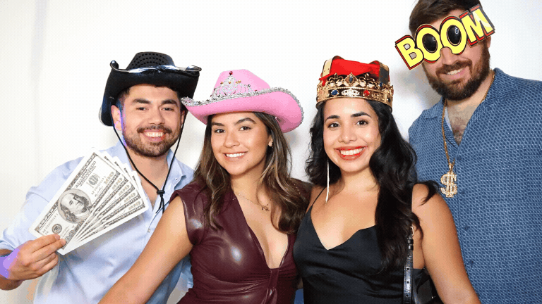 Group of four friends dressed in party costumes and accessories, smiling in front of a white background. One person wears a sheriff's hat and holds dollar bills, another wears a pink cowboy hat, the third has a crown and a black dress, and the fourth