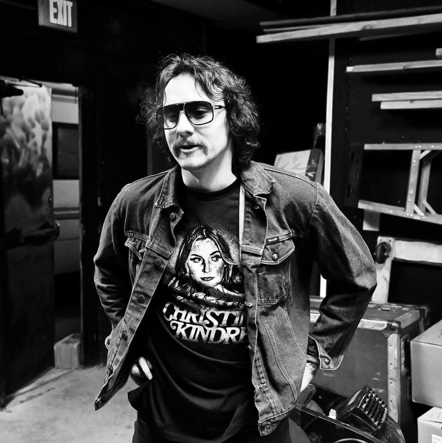 Isaac King Rudd TubeArk Amp Amplifier A man with curly hair wearing sunglasses, a graphic t-shirt, and a denim jacket stands in a cluttered room, possibly a backstage or storage area. Musician backstage