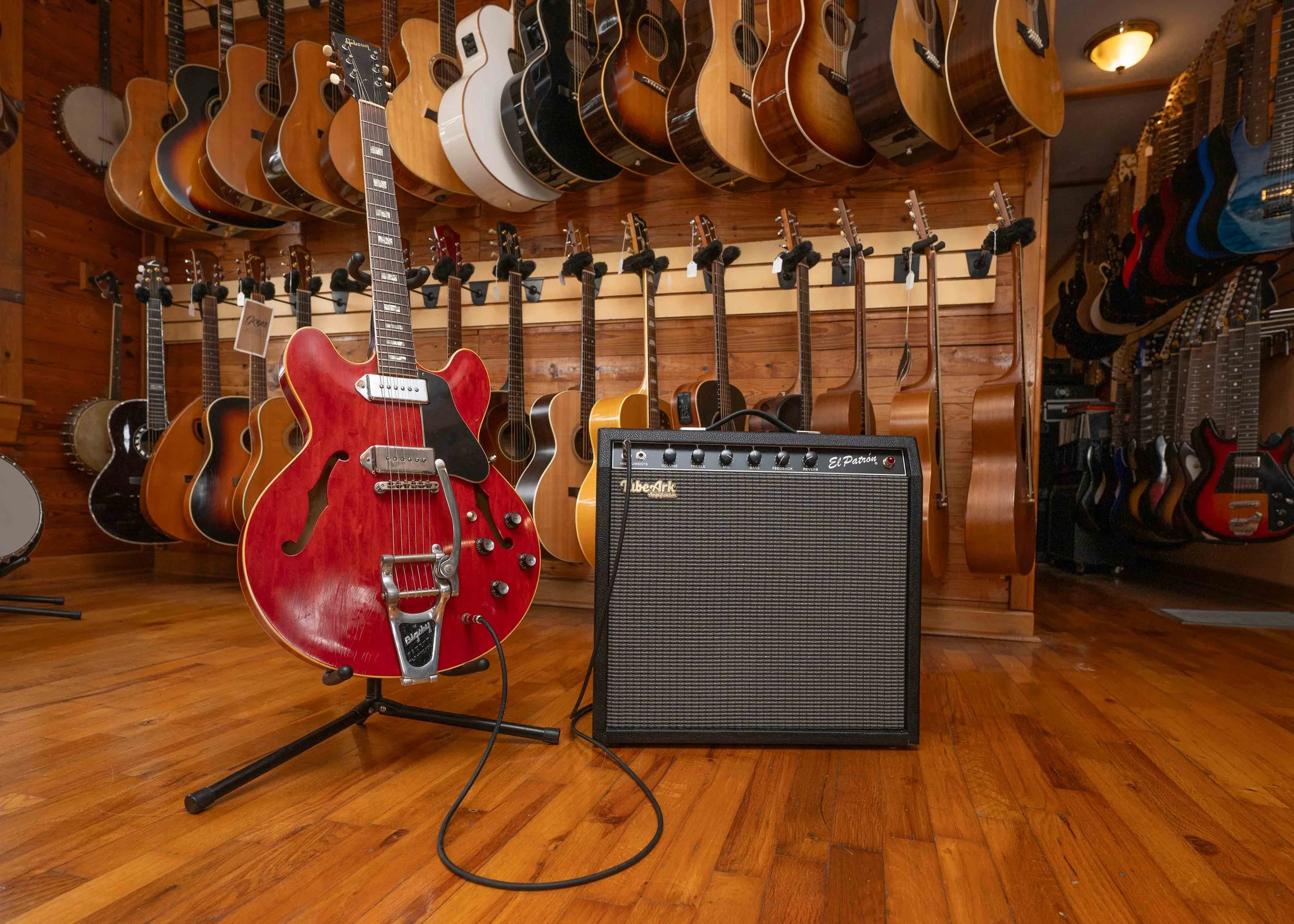 A display of various acoustic and electric guitars hanging on a wooden wall, including a red semi-hollow electric guitar in the foreground, with an amplifier and additional guitars on the floor and to the right.