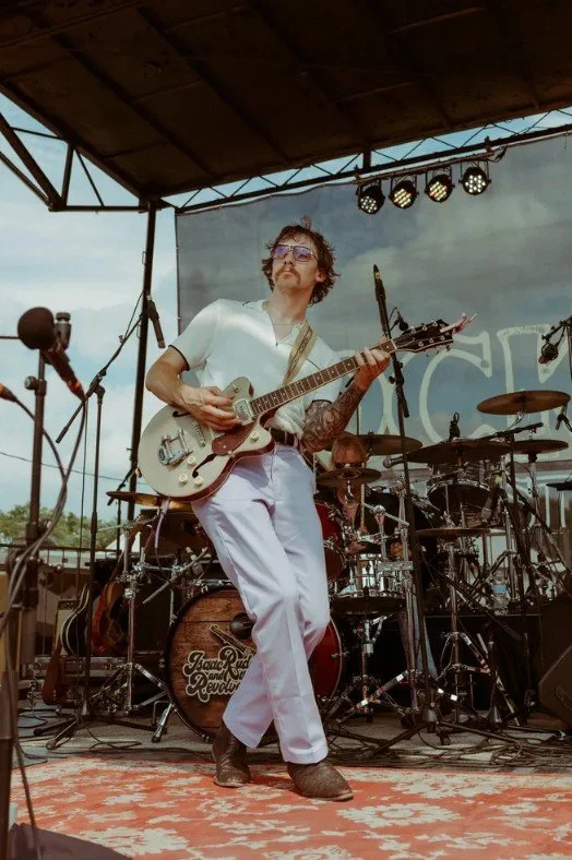 Musician playing electric guitar on outdoor stage with drum set and stage lights. Isaac King Rudd TubeArk Amplifier