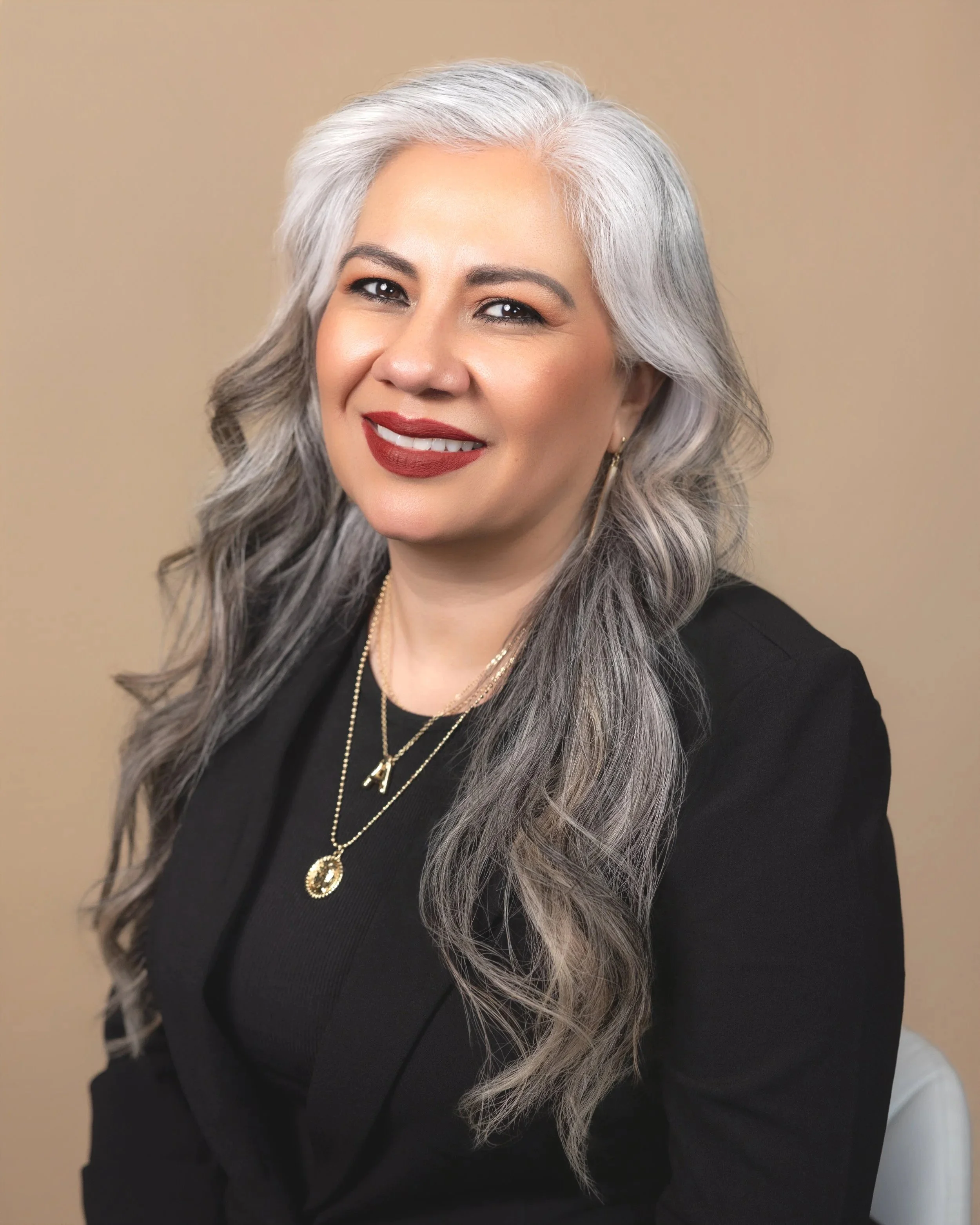 A woman with long, wavy gray hair, wearing red lipstick, a black top, and layered gold necklaces, smiling against a beige background.