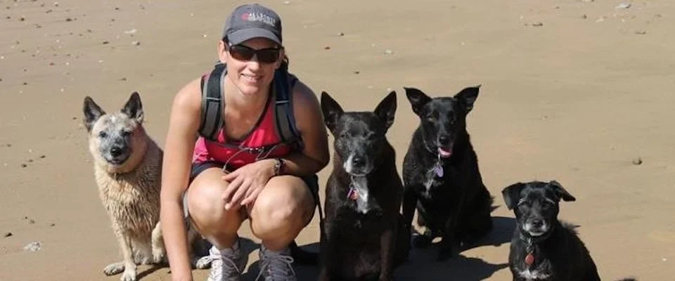 Dog trainer, Debbie Coleman, kneels on a sandy beach next to four dogs of various breeds.