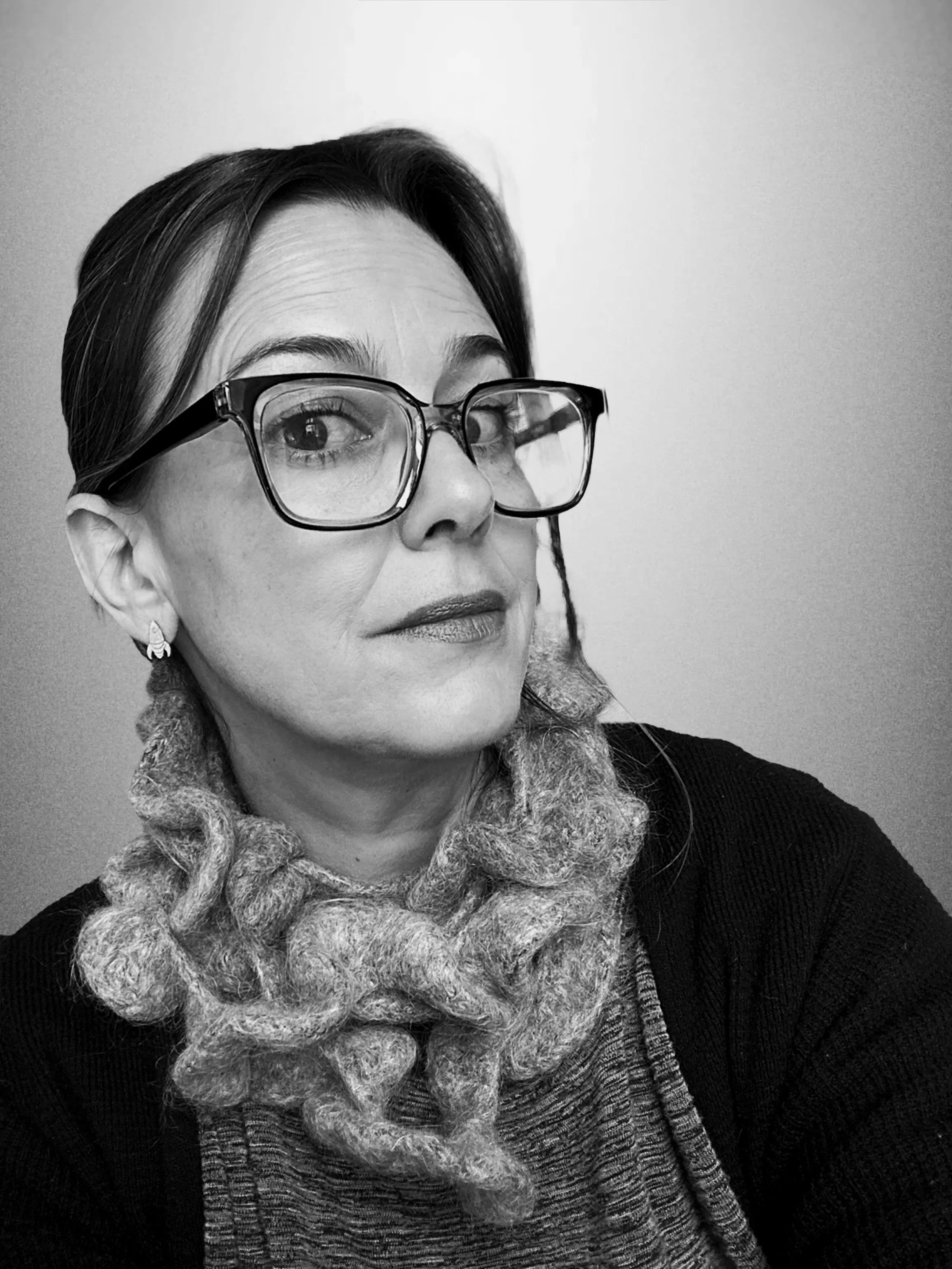 A black and white portrait of a woman with short hair, wearing large glasses, earrings, a knitted scarf, and a sweater, posing against a plain background.