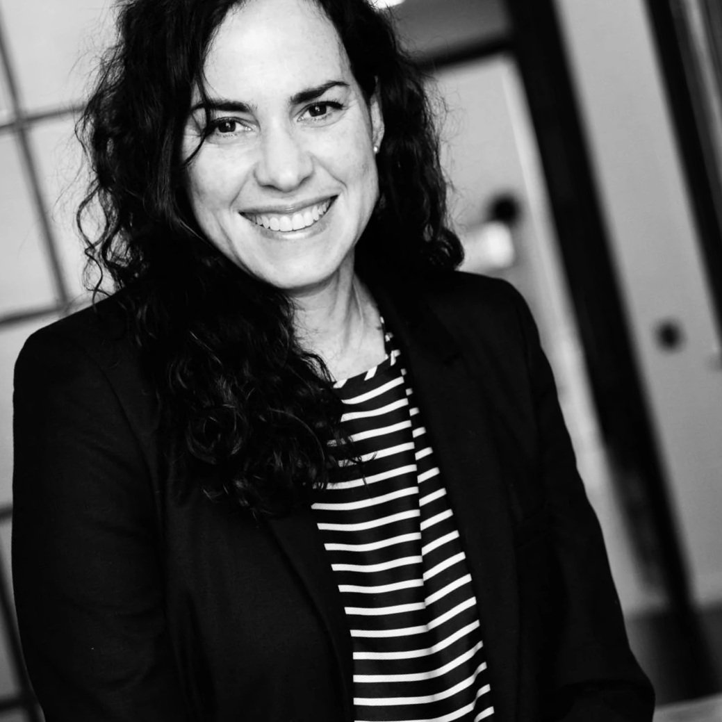 Black and white photo of a smiling woman with dark, curly hair, wearing a striped shirt and a dark blazer, standing indoors.