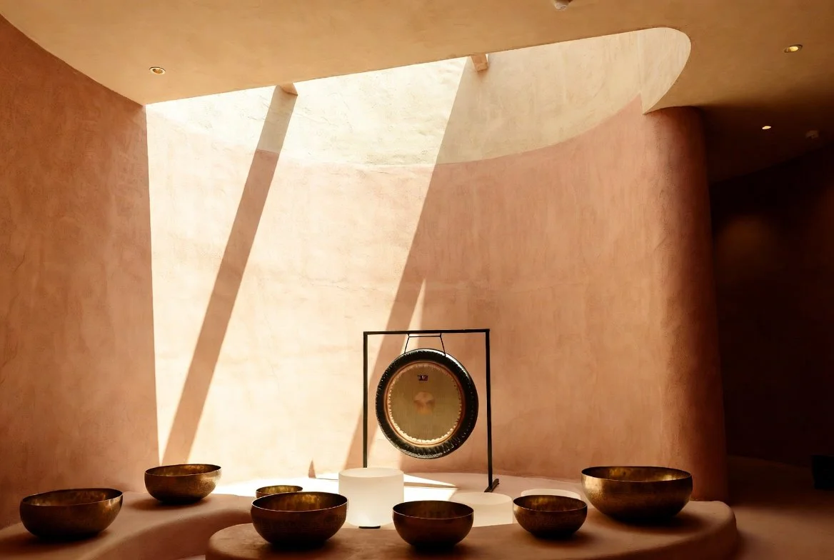 An interior with a sunlit wall, six bronze bowls arranged on a curved surface, and a gong hanging on a black stand in the center.