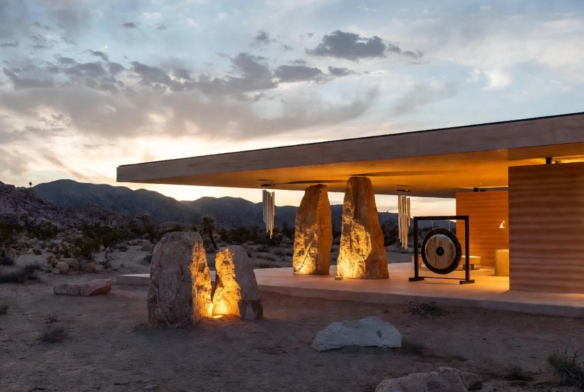 Modern desert house exterior during sunset with large stone sculptures and a gong, illuminated by warm lighting.