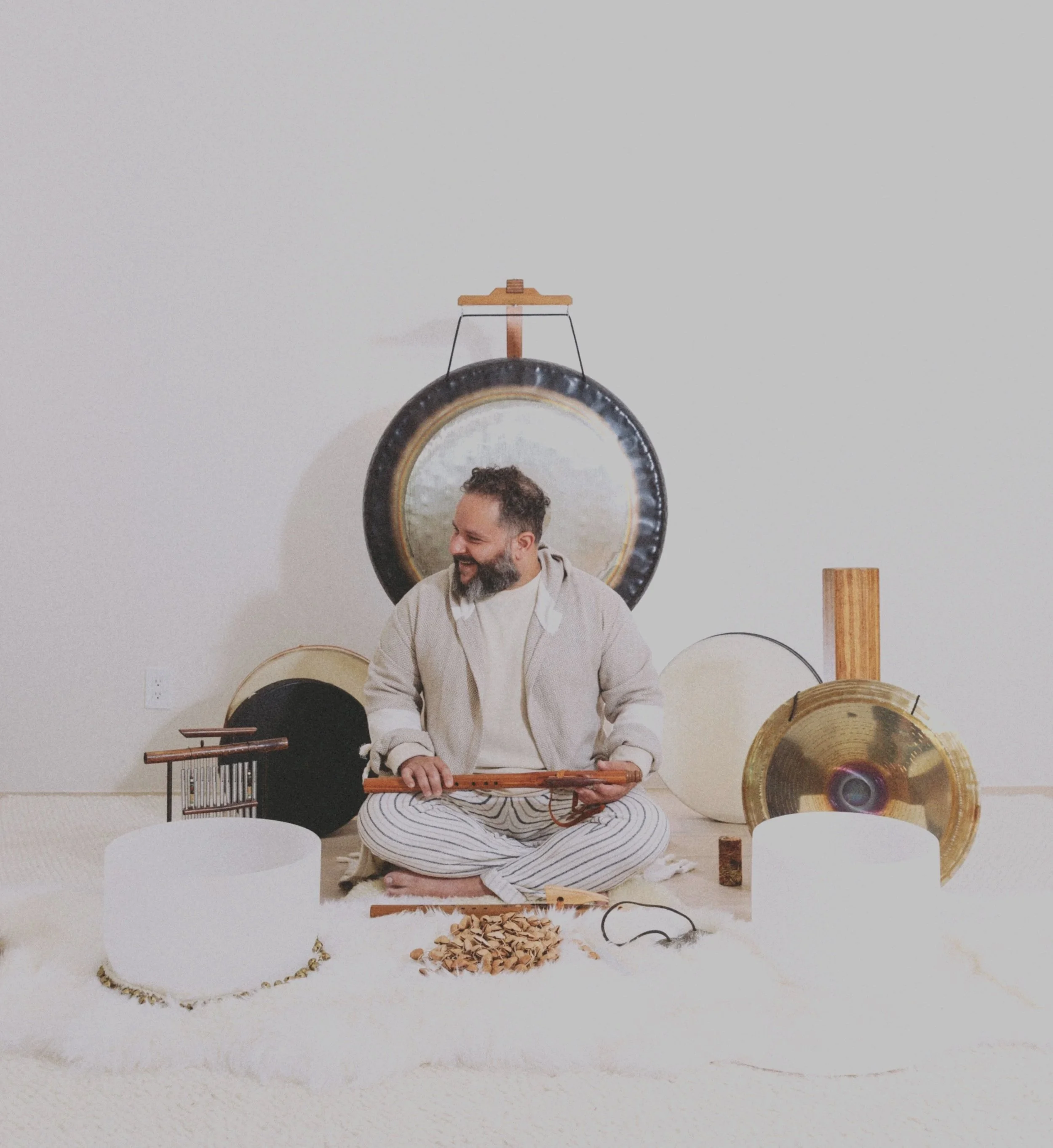 A man with a beard and short hair sits cross-legged on the floor playing a musical instrument, surrounded by musical objects like records and a xylophone, with a circular mirror on the wall behind him reflecting his face and various decorative items.