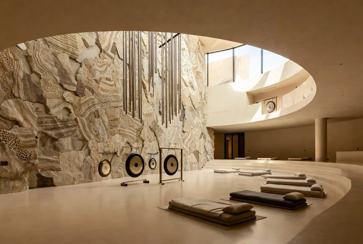 Interior of a modern meditation hall featuring a curved stone wall with textured mineral-like patterns, hanging gongs, lotus-shaped pads for seating, and large windows allowing natural light to illuminate the space.