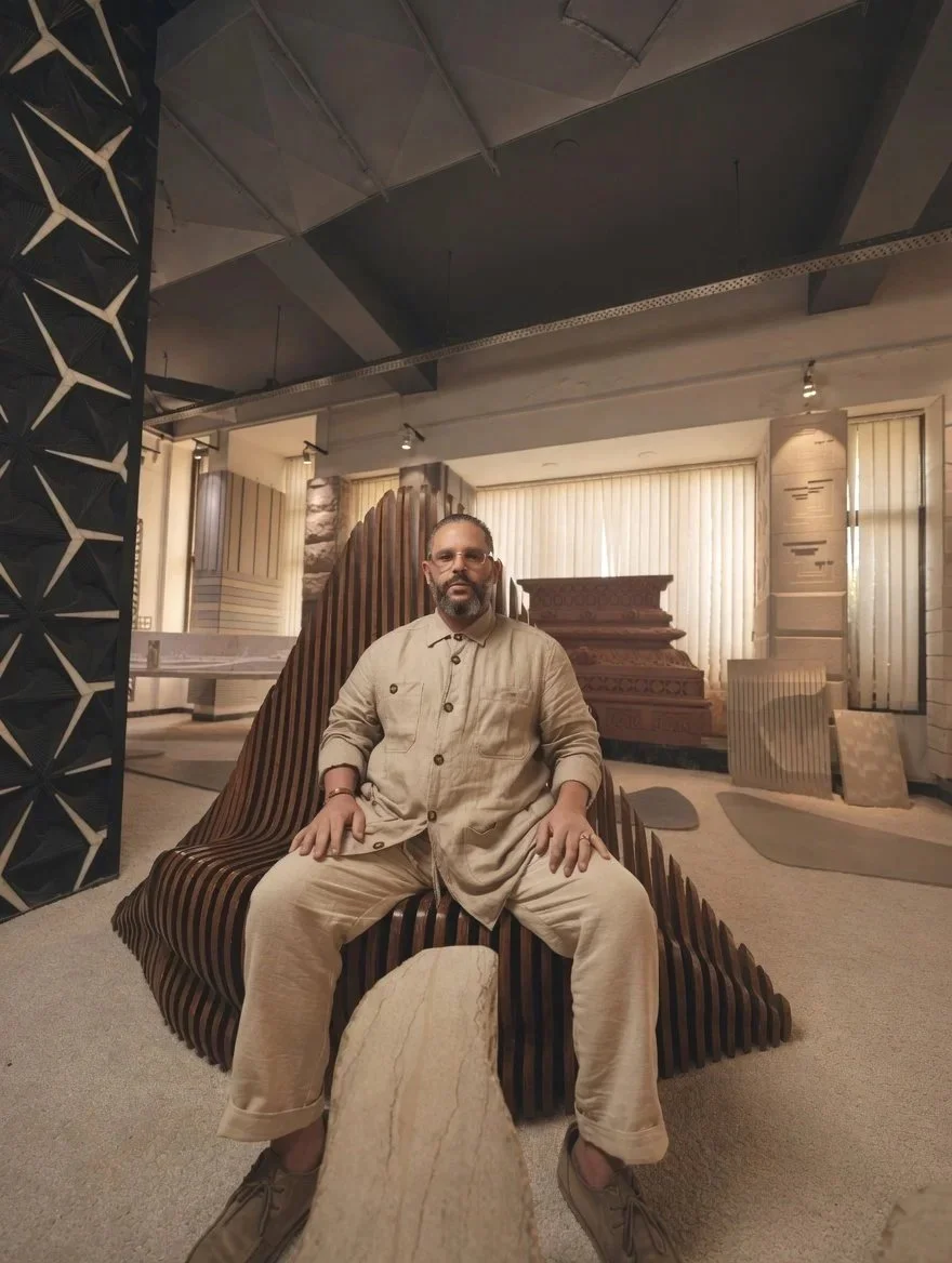 A man with a beard, glasses, and beige clothing sitting on a uniquely shaped wooden bench inside a modern, well-lit room with architectural decor.
