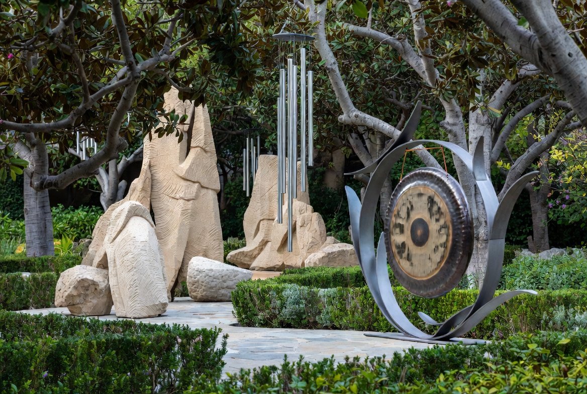 Outdoor garden scene featuring large beige stone sculptures, a metal wind chime with multiple tubes, and a metal sculpture with a round central element surrounded by curved metal pieces, set among green bushes and trees.