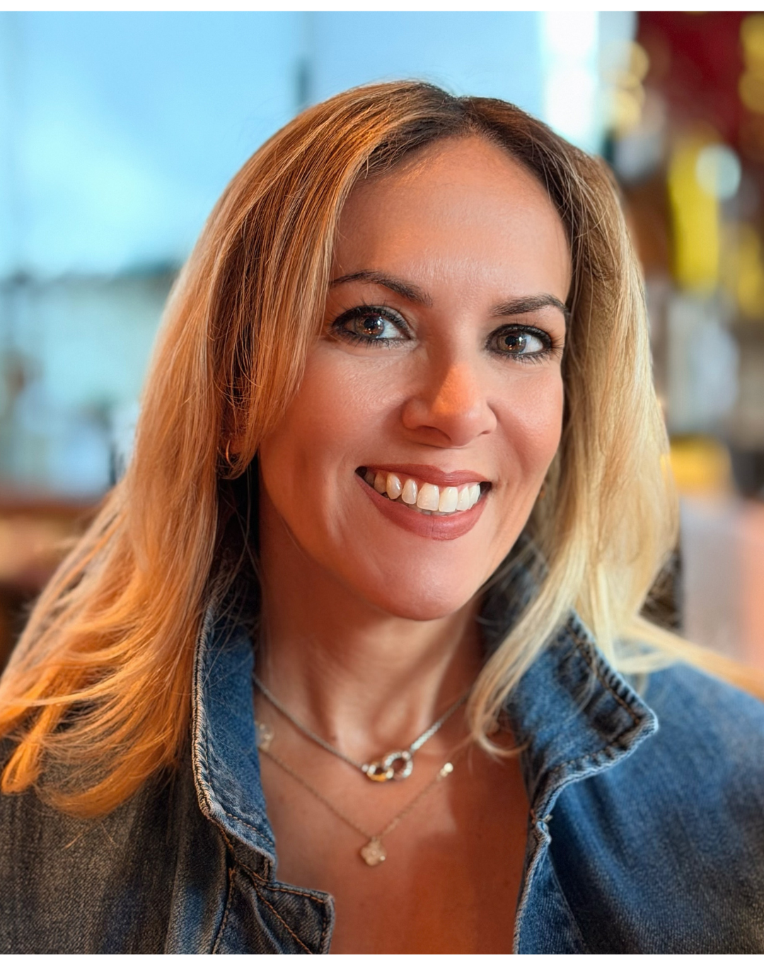 A smiling woman with blonde hair, wearing a denim jacket and layered necklaces, in a brightly lit indoor setting.