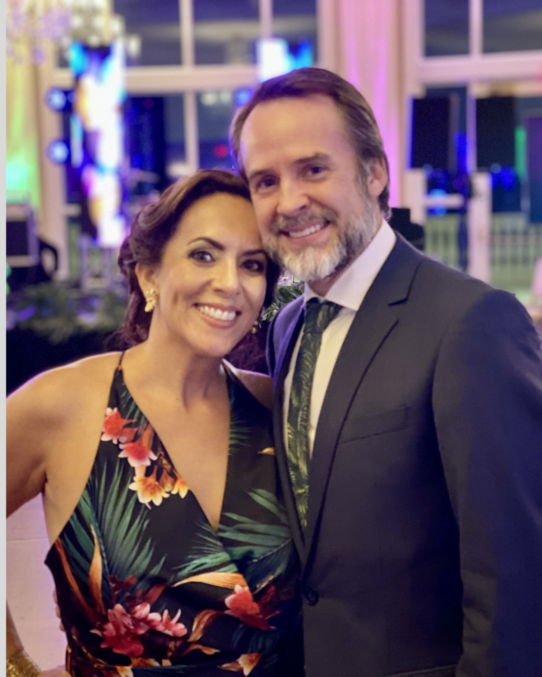 A smiling woman and man in formal attire at an indoor event, with colorful lighting and a blurred background.